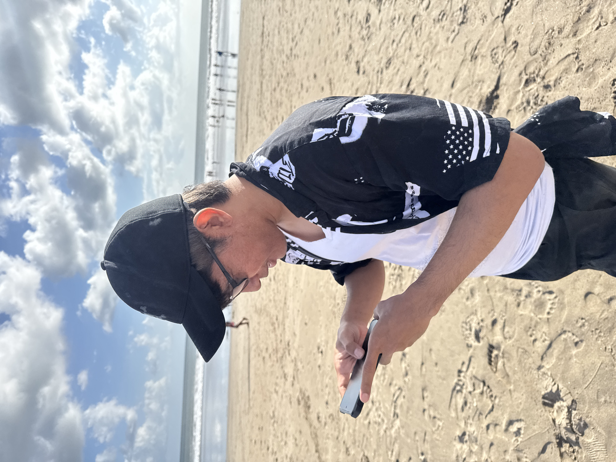 A young man, wearing a black baseball cap, glasses, a white tank top, and a black short-sleeved shirt with a white flag design, stands on the sandy beach in Kuta, Indonesia, looking down at his smartphone. The ocean is visible in the background, with gentle waves lapping the shore and several people in the distance. The sky is partly cloudy with a mix of blue sky and white clouds. The sand is light brown and shows footprints from people walking on the beach. The scene is bathed in natural daylight, suggesting it is daytime.
