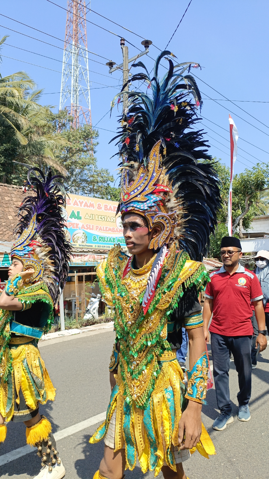 In Krajan Lor, Indonesia, two individuals in elaborate traditional attire participate in a parade. The person in the foreground, facing right, wears a tall headdress adorned with black and blue feathers, intricate gold and turquoise embellishments, and red accents. Their costume features a gold and green sequined chest piece, a layered gold and turquoise skirt, and turquoise armbands. They have face paint and appear to be walking. To the left, another person in similar, though less elaborate, attire is also visible. In the background, other parade participants and onlookers are present, including a man in a red shirt and dark pants, and a person in a white mask. A telecommunications tower stands tall against a clear blue sky, and a billboard for "AYAM & BAKSO AJI LESTARI PEK ASLI PALEMBANG" is visible. The scene takes place on a paved road under bright sunlight.