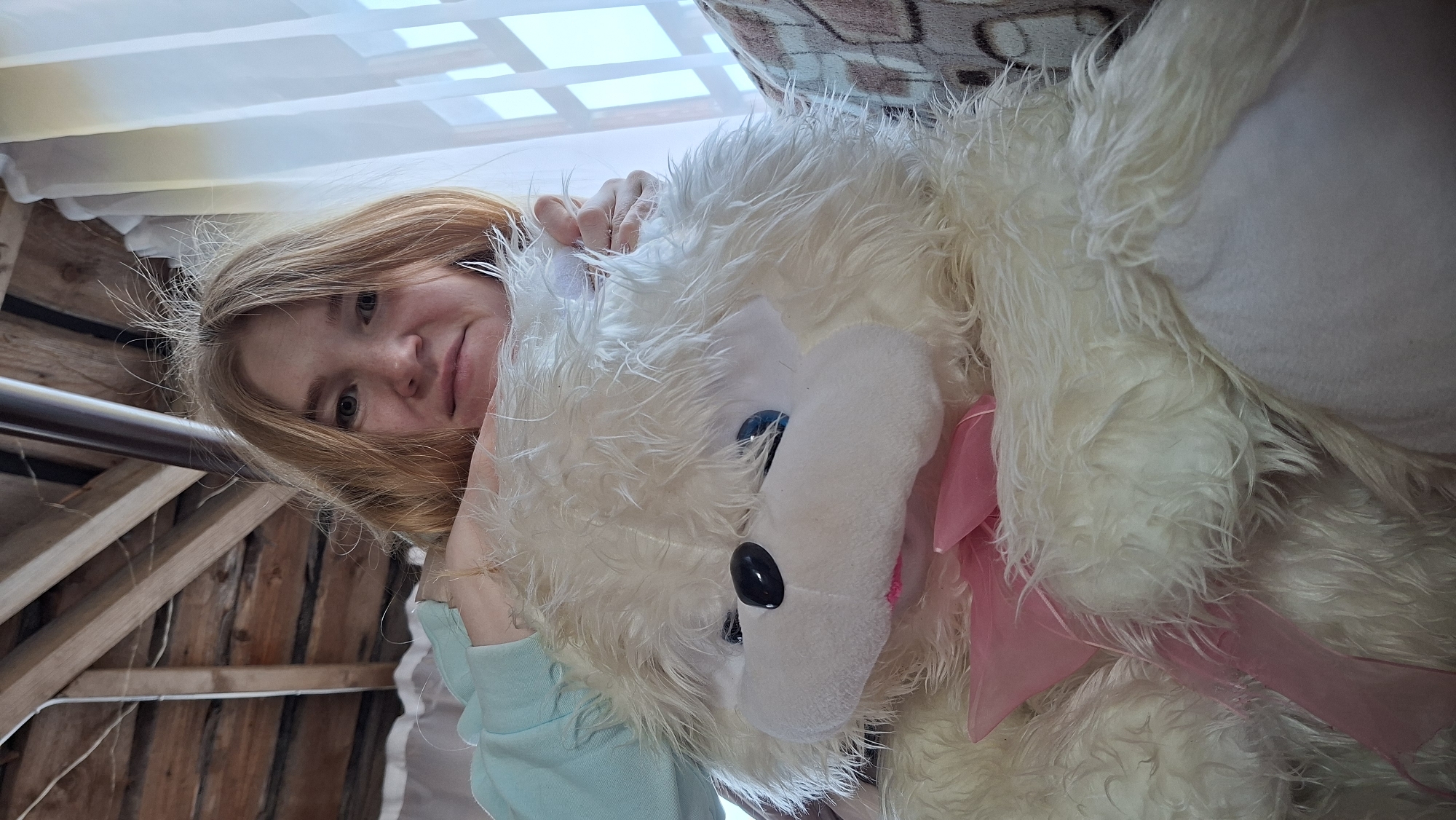 A fair-skinned, blonde-haired young woman is posing with a large, white, fluffy stuffed animal with a pink ribbon around its neck. The woman is smiling. The background shows a wooden structure, possibly an attic, with a window covered by a white curtain. The location is possibly in Большой Букор, Russia.