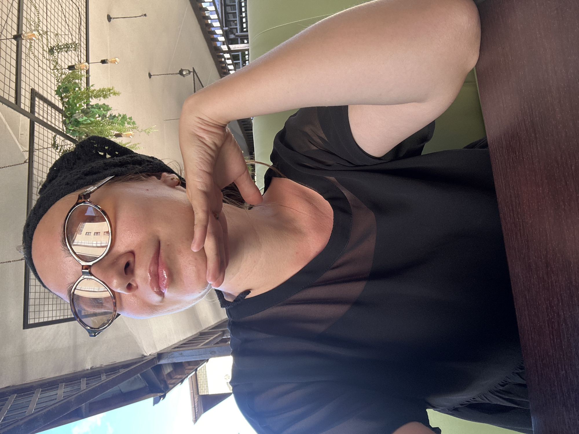 A middle-aged Caucasian woman is pictured from the chest up, wearing a black knitted beanie and large, tortoiseshell-framed glasses. She has on a sheer black top with delicate ruffles at the neckline. Her right hand is resting on her chin, and her left arm is visible in the foreground, bent at the elbow. She is seated at a dark wood table, with a green booth visible behind her. The background suggests an outdoor covered dining area with a lattice ceiling, decorative plants, and light fixtures. The lighting is bright, indicating daytime. The overall scene is relaxed and casual. The image was taken in Vityazevo, Russia.