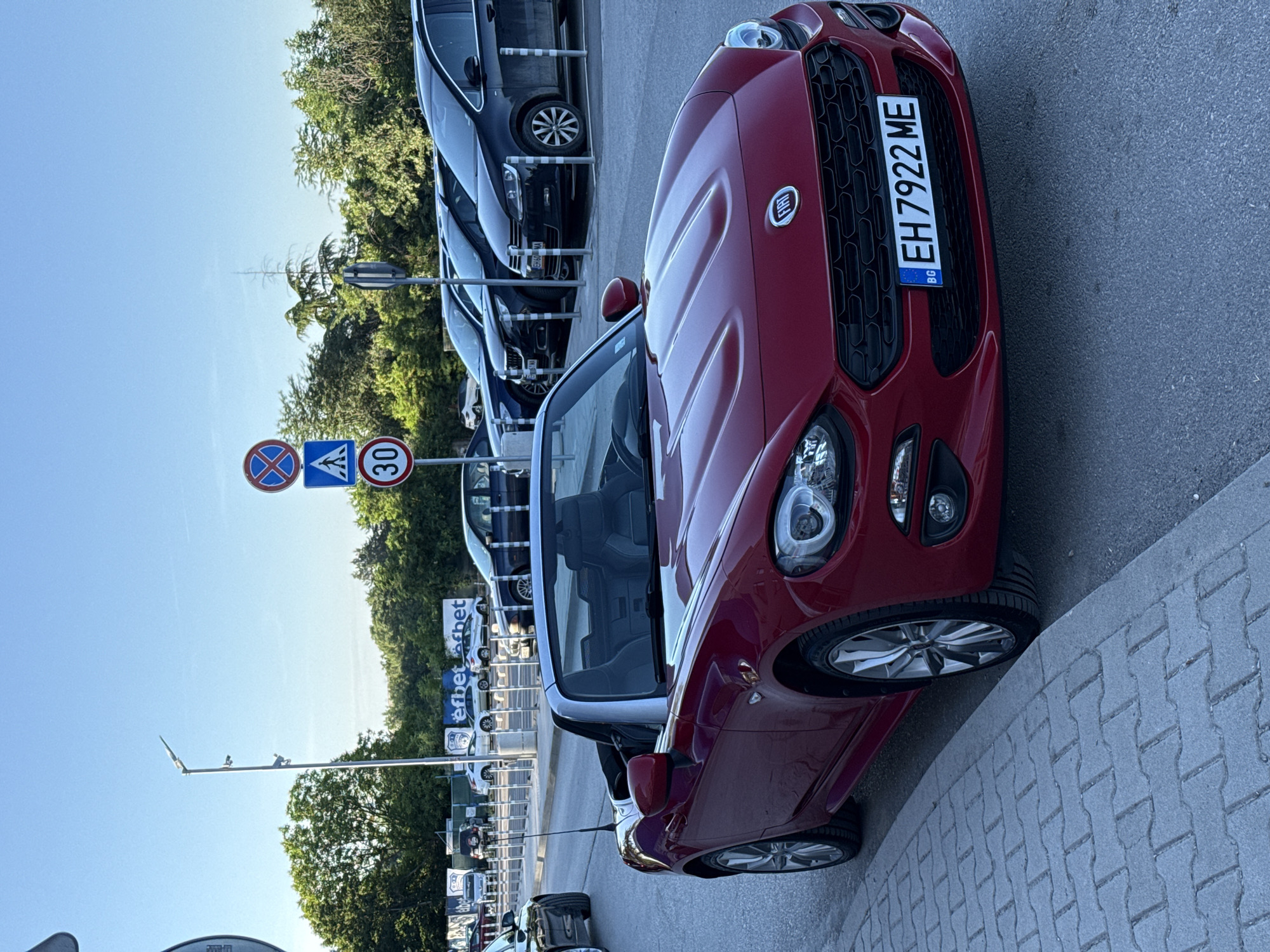 The image shows a red convertible Fiat 124 Spider parked on a street in Pleven, Bulgaria. The car is positioned diagonally, facing the viewer, with its front end slightly angled towards the right. The vehicle has a silver fabric soft top that is up. The license plate is visible and reads "EH 7922 ME". The car is parked next to a sidewalk made of interlocking pavers, and behind it is a row of other parked cars, including black sedans and a minivan, in a parking lot. In the background, there are trees and some buildings with advertising banners. Traffic signs are visible on the right side of the image, including a "no stopping" sign, a pedestrian crossing sign, and a "30" speed limit sign. The sky is clear and blue, indicating it is daytime. There are no people visible in the image.