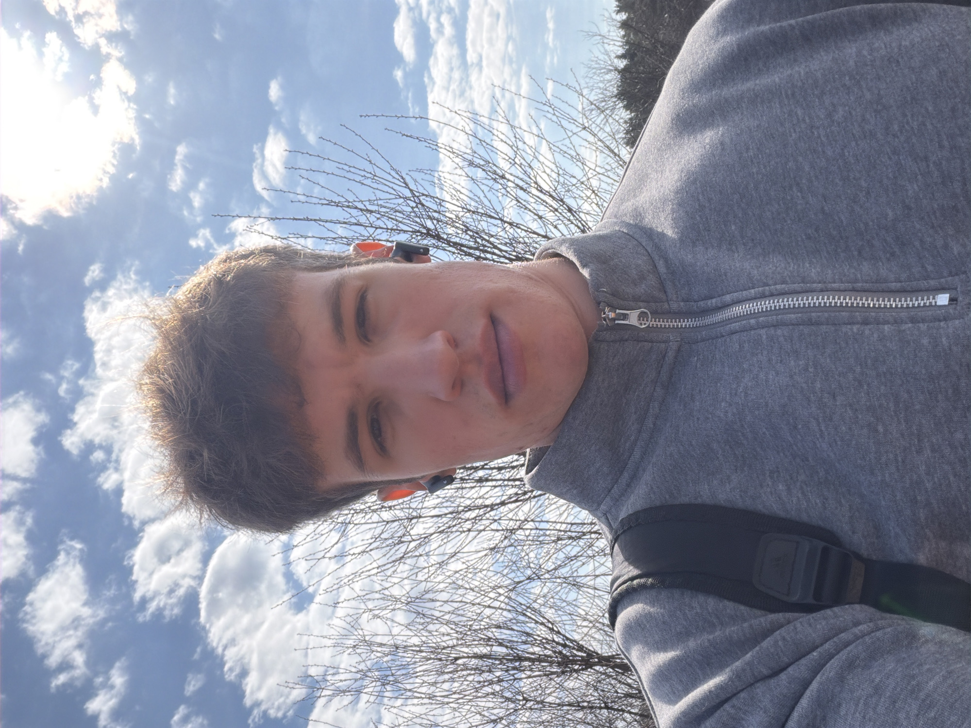 A young man is featured in a close-up, low-angle selfie, looking directly at the camera with a neutral expression. He has short, light brown hair. He is wearing a grey zippered jacket, and a black backpack strap is visible over his right shoulder. Black and orange wireless earbuds are visible in both ears.
The background shows a bright blue sky with scattered white, fluffy clouds. The sun is partially visible in the upper left, casting a strong, bright light. Leafless tree branches, suggesting late autumn, winter, or early spring, are visible against the sky. The scene is outdoors, indicating bright daytime conditions in Прикамский, Russia. There is no visible text.