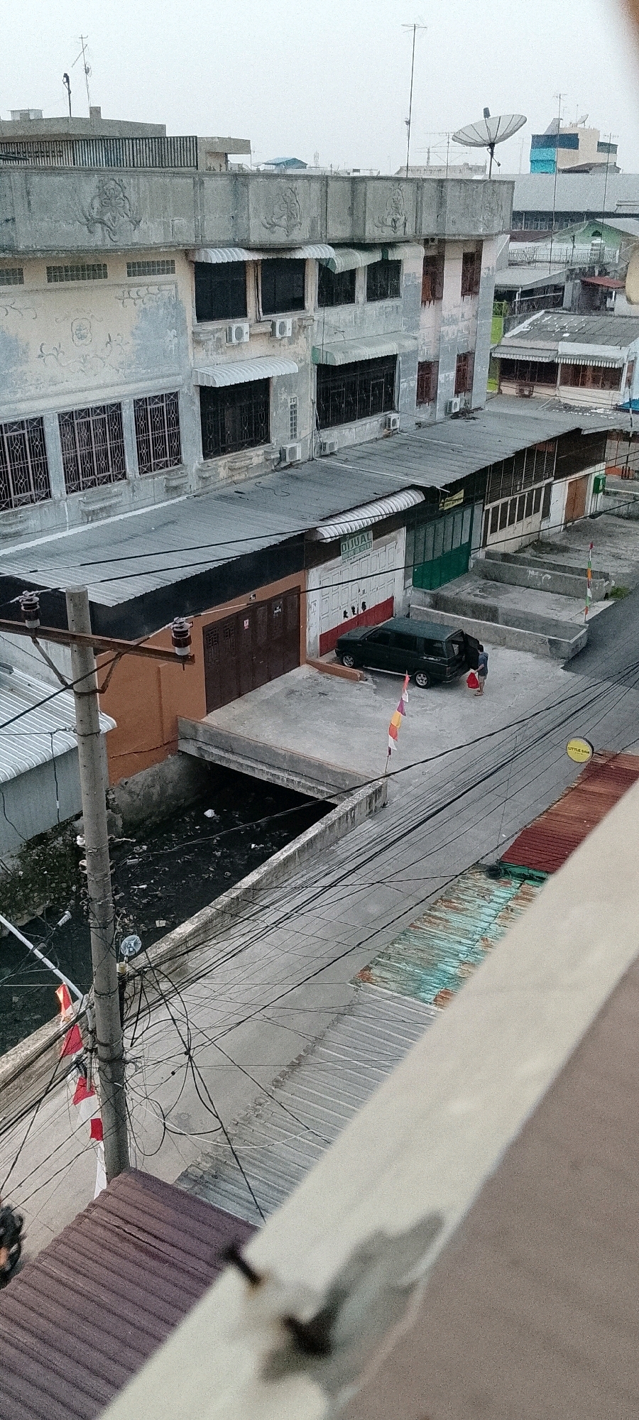 The image shows a street scene in the city of Medan, Indonesia, with a focus on a commercial area and an adjacent drainage canal. The buildings are a mix of older, multi-story structures and single-story storefronts, some appearing weathered and in need of repair. A dark-colored SUV is parked in front of one of the storefronts, with its rear hatch open. A person is standing next to the open hatch, holding a red bag. Several flags, red and white, are visible, attached to poles along the street and near the canal. The drainage canal is filled with dark, murky water and debris, indicating poor sanitation. Overhead, a complex network of electrical wires and cables crisscrosses the scene, suspended from utility poles. The sky is overcast, suggesting a muted light. In the background, other buildings and structures are visible, typical of an urban environment.