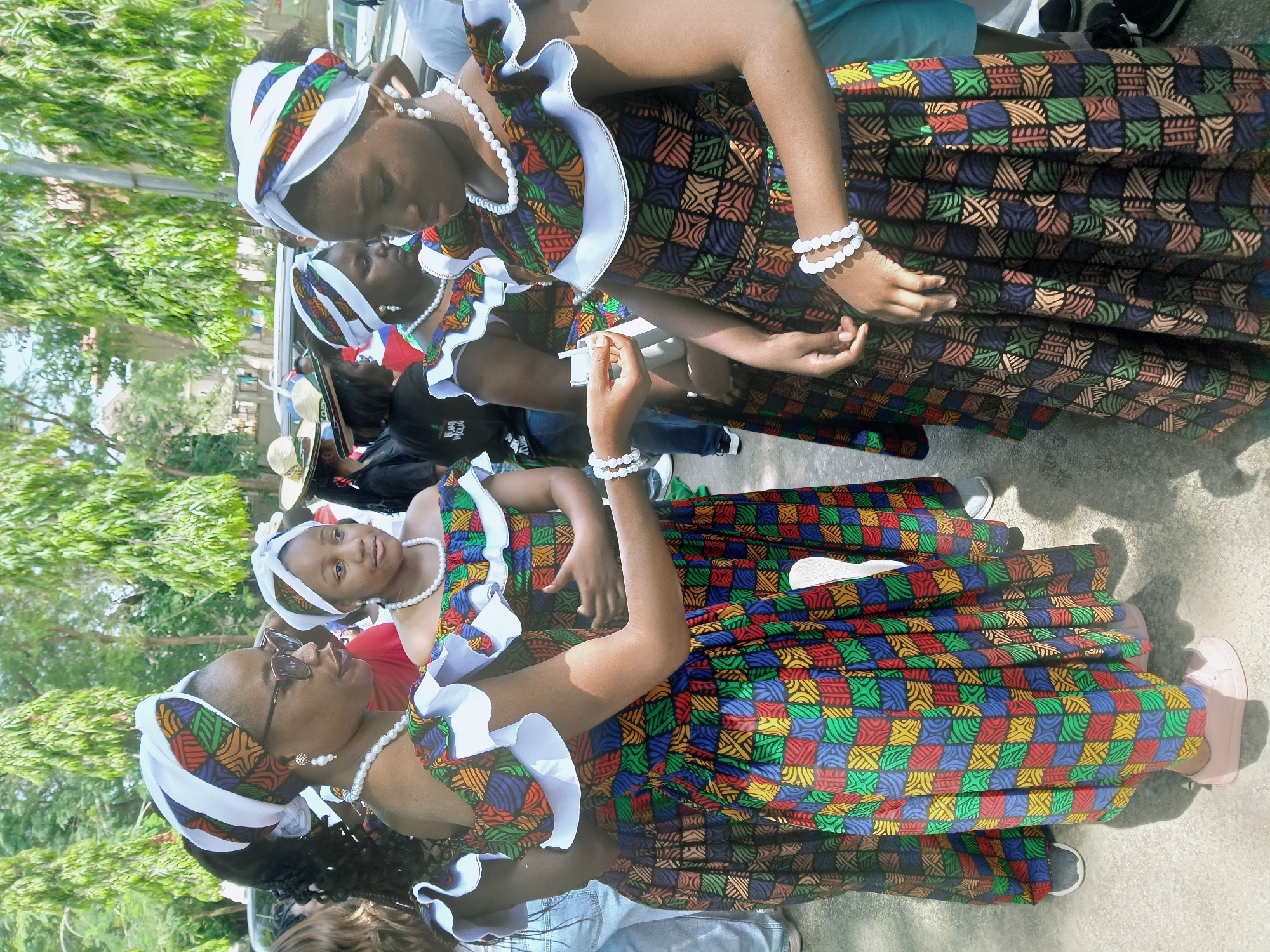Four Black women are wearing patterned dresses and headwraps. They are outside in an urban environment with green trees in the background. The location is Abuja, Nigeria. The women are adorned with white necklaces and bracelets. One woman is wearing sunglasses. Several people can be seen in the background, partially obscured.