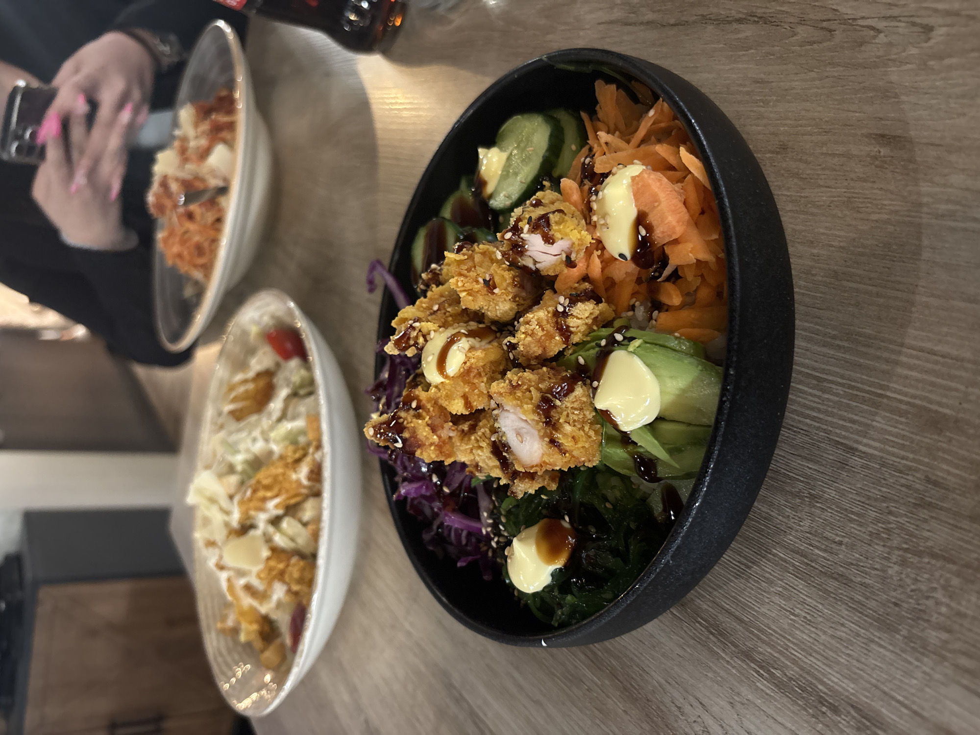 The image captures a dining scene featuring three bowls of food on a wooden-textured surface. The primary focus is a black bowl filled with a colorful assortment of ingredients: crispy fried chicken, seaweed, shredded carrots, sliced cucumbers, avocado, and shredded red cabbage. These elements are topped with dollops of a creamy sauce and a drizzle of a dark sauce, possibly teriyaki.
To the left of the primary bowl is another white bowl containing a similar composition, but with a different arrangement and balance of ingredients.
In the background, partially visible, is a third bowl and a person's hand holding a phone. A beverage bottle is also present in the upper background.
The setting appears to be a casual dining environment, possibly a restaurant. The location details, the city, and additional context are unknown.