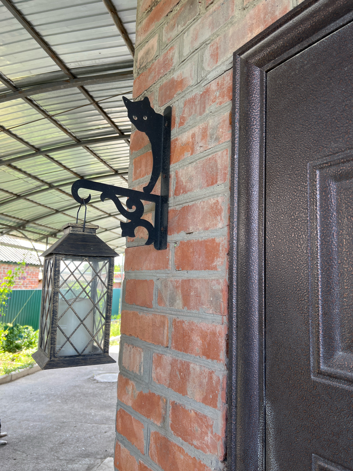 The image shows a close-up view of a brick wall with a black metal door frame and a lantern hanging from a bracket. The bracket is in the shape of a cat, with its head and front paws visible. The lantern is old-fashioned, with glass panels and a candle inside. The brick wall is made of red bricks and has some mortar showing. The door frame is dark brown and appears to be made of metal. In the background, a corrugated metal roof is visible, suggesting an outdoor covered area. The overall scene appears to be the entrance to a building, possibly a house or a shop. There are no people visible in the image. The location is identified as Morozovsk, Russia.
