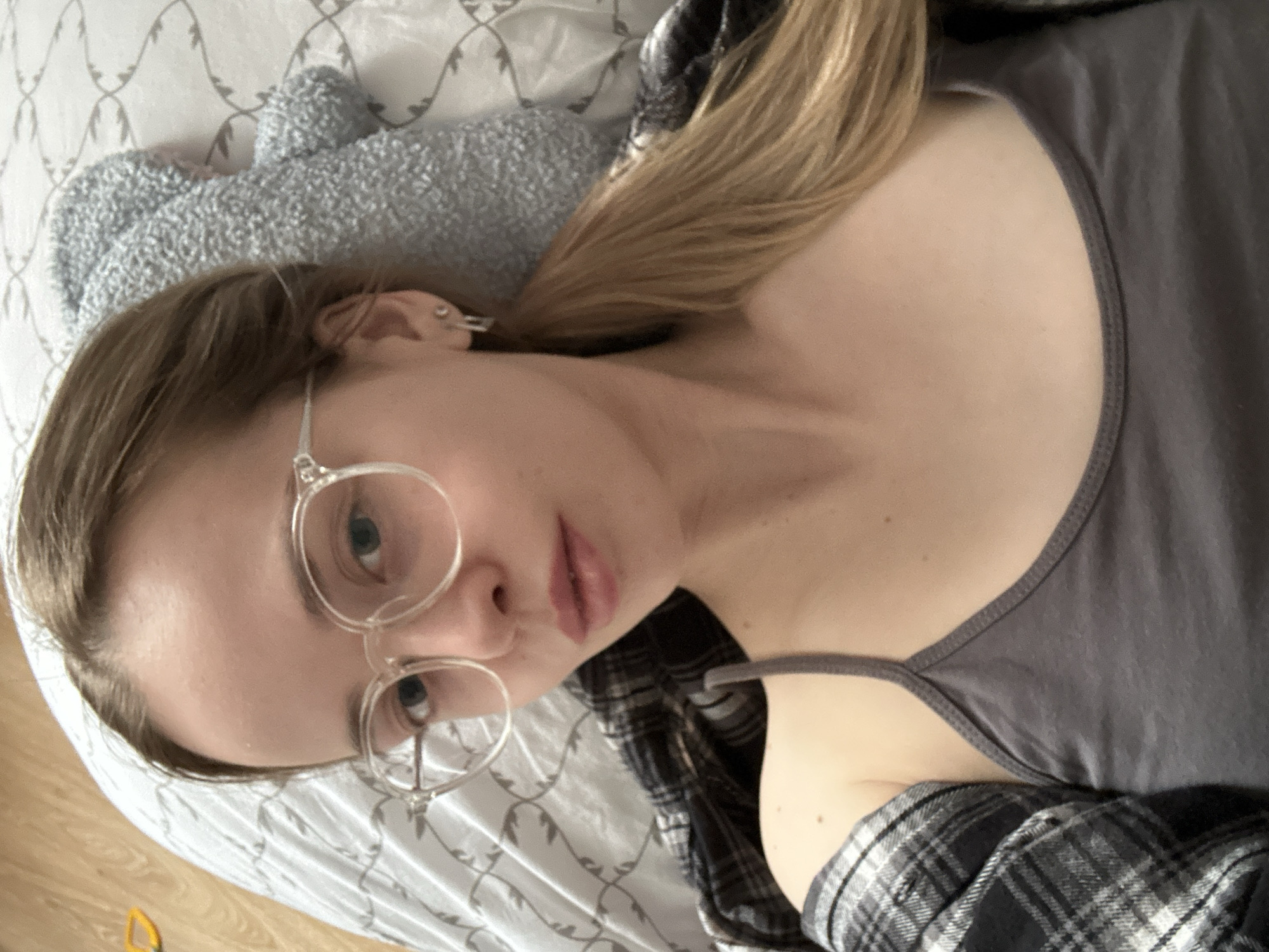 A woman with blonde hair and round glasses lies on a bed. She wears a gray tank top and a black and white plaid shirt. A gray, plush object is visible near her head on a white bedsheet patterned with gray designs. The location is not discernible.