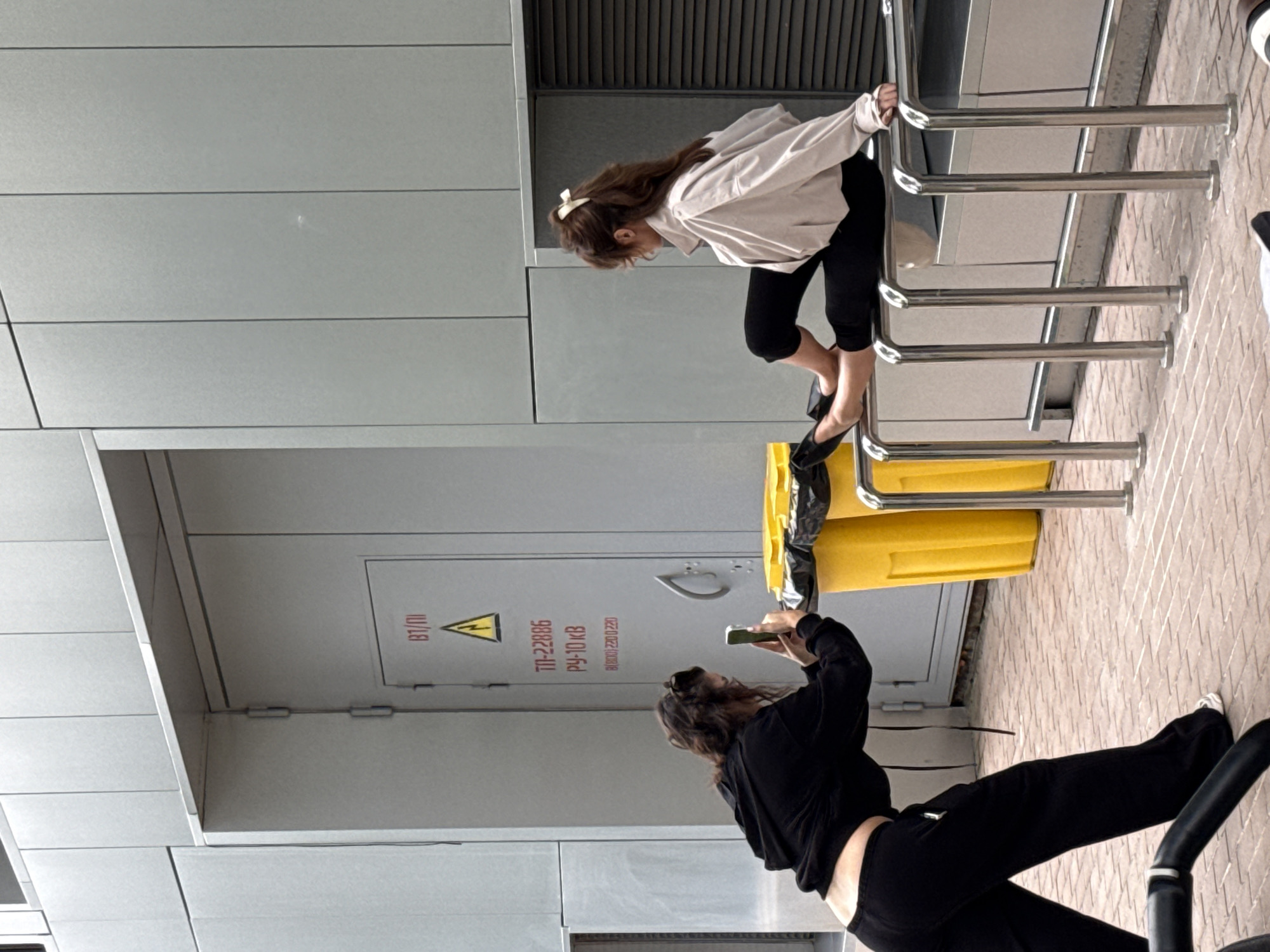 The image shows two young women in an urban outdoor setting, possibly a public space like a park or plaza in Moscow, Russia. The scene is captured from a slightly elevated perspective, and the image is oriented vertically.
In the foreground, one woman is captured from the waist up, wearing a black hooded sweatshirt and black pants. She is holding a smartphone in her right hand, appearing to take a photo or video. Her head is tilted upwards, and her hair is casually styled.
In the background, another woman is positioned on a set of metal railings, which appear to be part of a bike rack or a similar public utility. She is wearing a light-colored long-sleeved shirt and black leggings. Her body is contorted as she balances on the railings, with her feet bare or wearing minimal footwear. She has a white bow in her hair. A bright yellow trash bin is visible behind her, with a black trash bag partially covering its opening.
The setting features modern architectural elements with large, flat, gray panels, suggesting a contemporary urban environment. There is also a brick-like patterned surface visible on the ground.
Text elements are visible on a gray panel behind the woman with the trash bin. One section shows a warning symbol (a triangle with an exclamation mark) and Cyrillic text, which when deciphered appears to be "TN-22886" and "РУ-10 KB". Other text includes what appears to be a phone number or code "8(000) 228-0228" and "IU/LEI".
The lighting suggests it is daytime, but the specific time is not clearly discernible. The overall mood of the image is candid and casual, capturing everyday activities in a public space.
