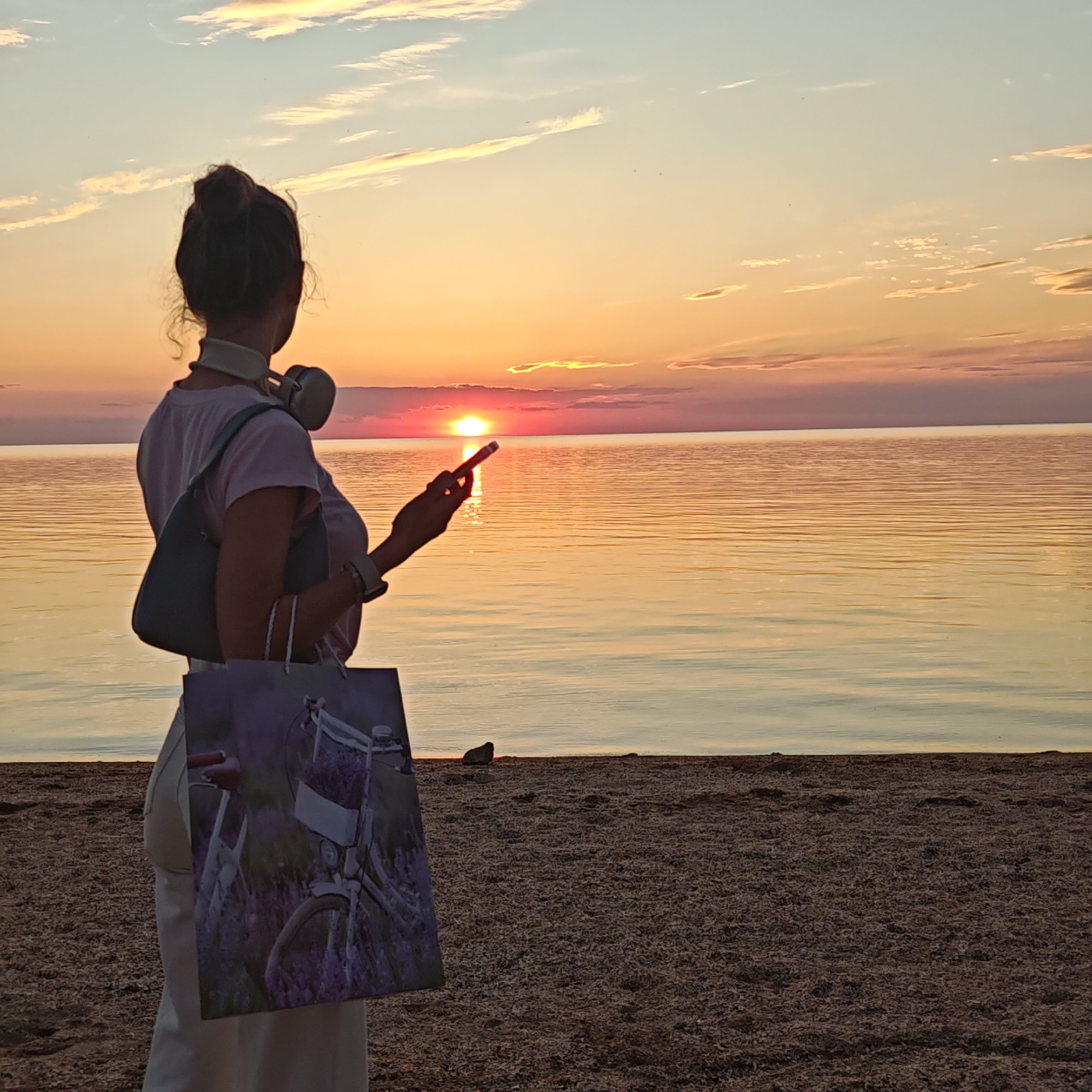 The image depicts a single individual standing on a sandy or gravelly beach at sunset, looking towards a large body of water. The location is in the city of Unknown, Unknown. The sun, a bright orange-red orb, is visible low on the horizon, partially obscured by clouds, casting warm hues across the sky and reflecting on the calm water.
The person, seen from behind, appears to be an adult with dark hair tied in a bun. She is wearing a light-colored short-sleeved top, a light-colored neck pillow, and light-colored over-ear headphones around her neck. A dark-colored shoulder bag is on her left shoulder. She holds a smartphone in her right hand, pointing it towards the setting sun, with its screen faintly illuminated. In her left hand, she carries a rectangular shopping bag displaying an image of a white bicycle with a basket of purple flowers. The foreground consists of a densely packed surface of small, dark granular material, likely shells or pebbles. The water extends to the horizon, meeting a sky with scattered light clouds tinted orange, pink, and pale blue. A small, dark object is visible on the waterline to the right of the person.