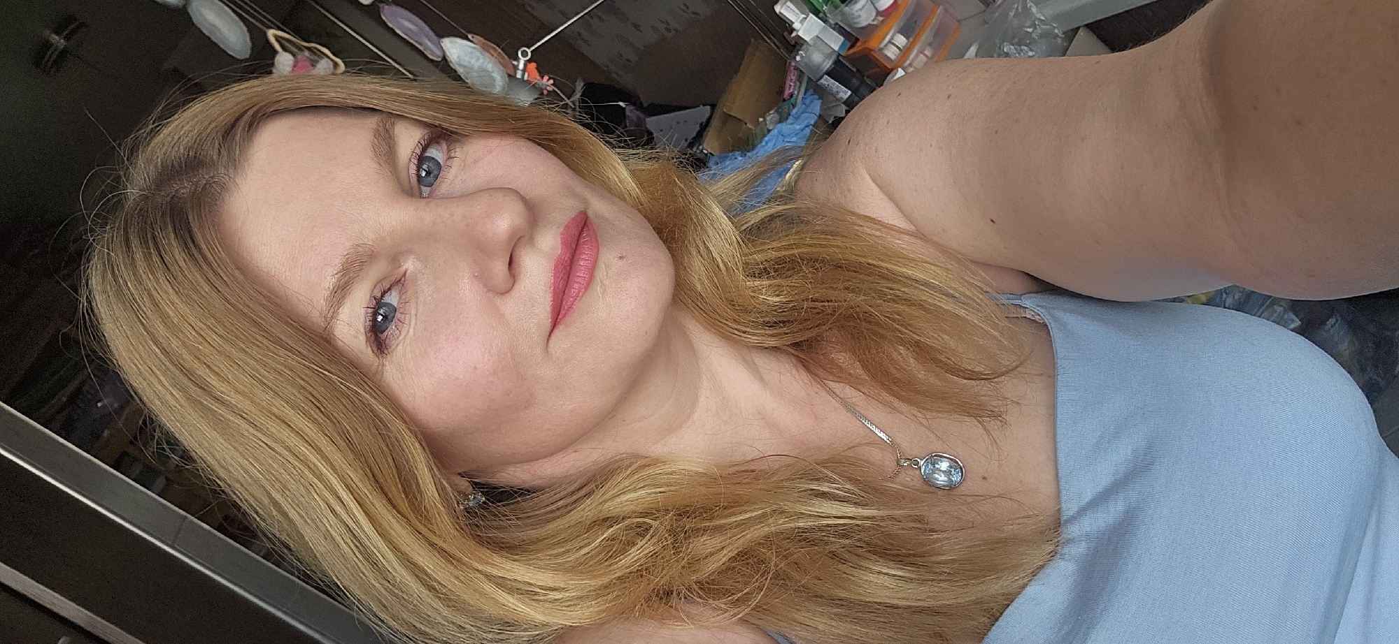 A close-up, overhead selfie of a woman with shoulder-length, wavy blonde hair and blue eyes. She is wearing a light blue tank top and a silver necklace with a light blue oval pendant. Her lips are painted a matte pink. The background is blurred and appears to be an indoor space with shelves and various objects, suggesting a home environment. There are no visible indications of her location in Krasnodar, Russia.