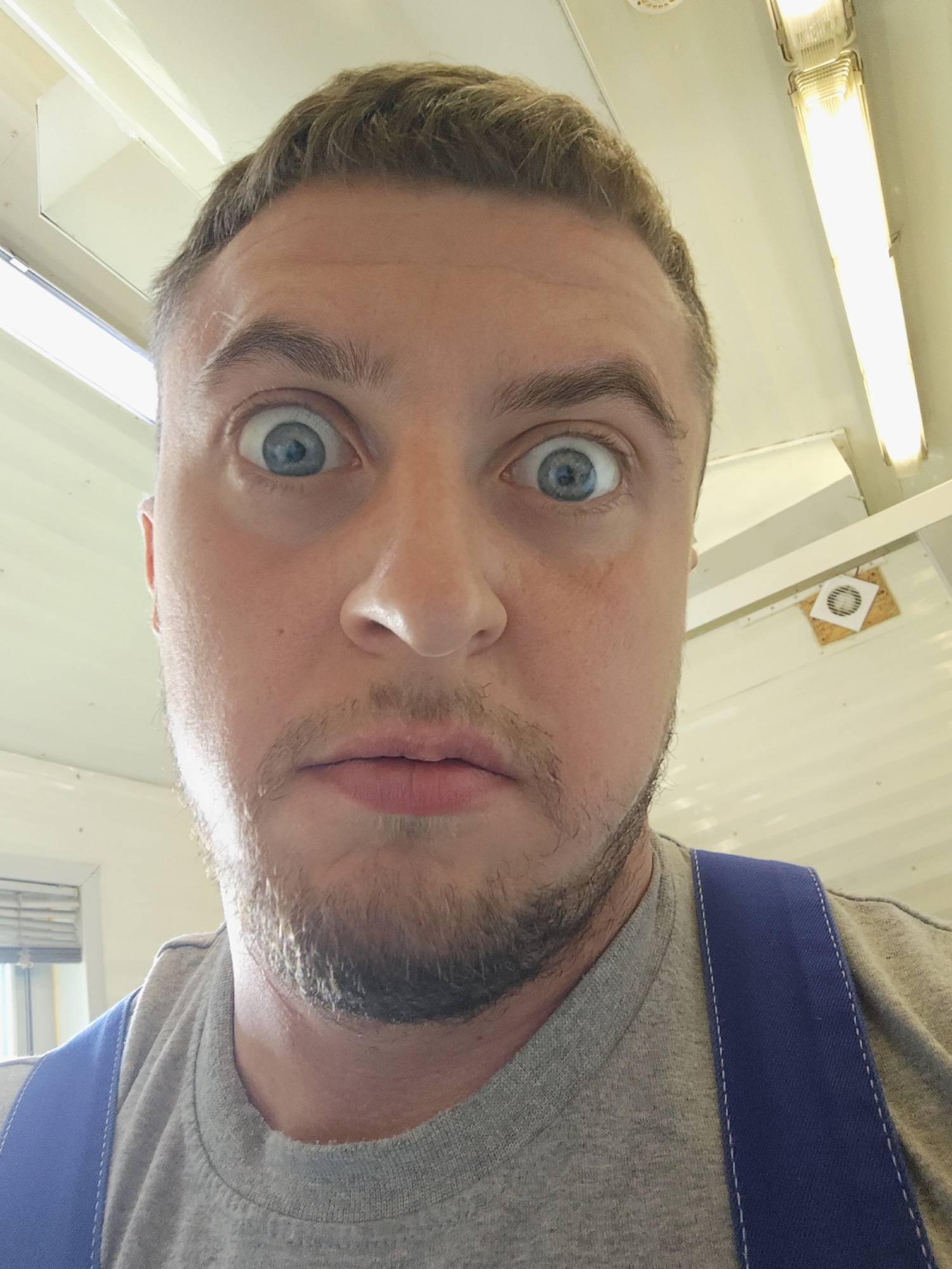 A close-up, slightly downward-angled portrait captures a man with light skin, light brown hair, and blue eyes exhibiting a wide-eyed, surprised expression. He has a beard and is wearing a grey t-shirt with blue suspenders. The background reveals a white, paneled ceiling with fluorescent lighting fixtures. A circular vent with a square paper attached to it is also visible on the ceiling. The location is Unknown, Unknown.