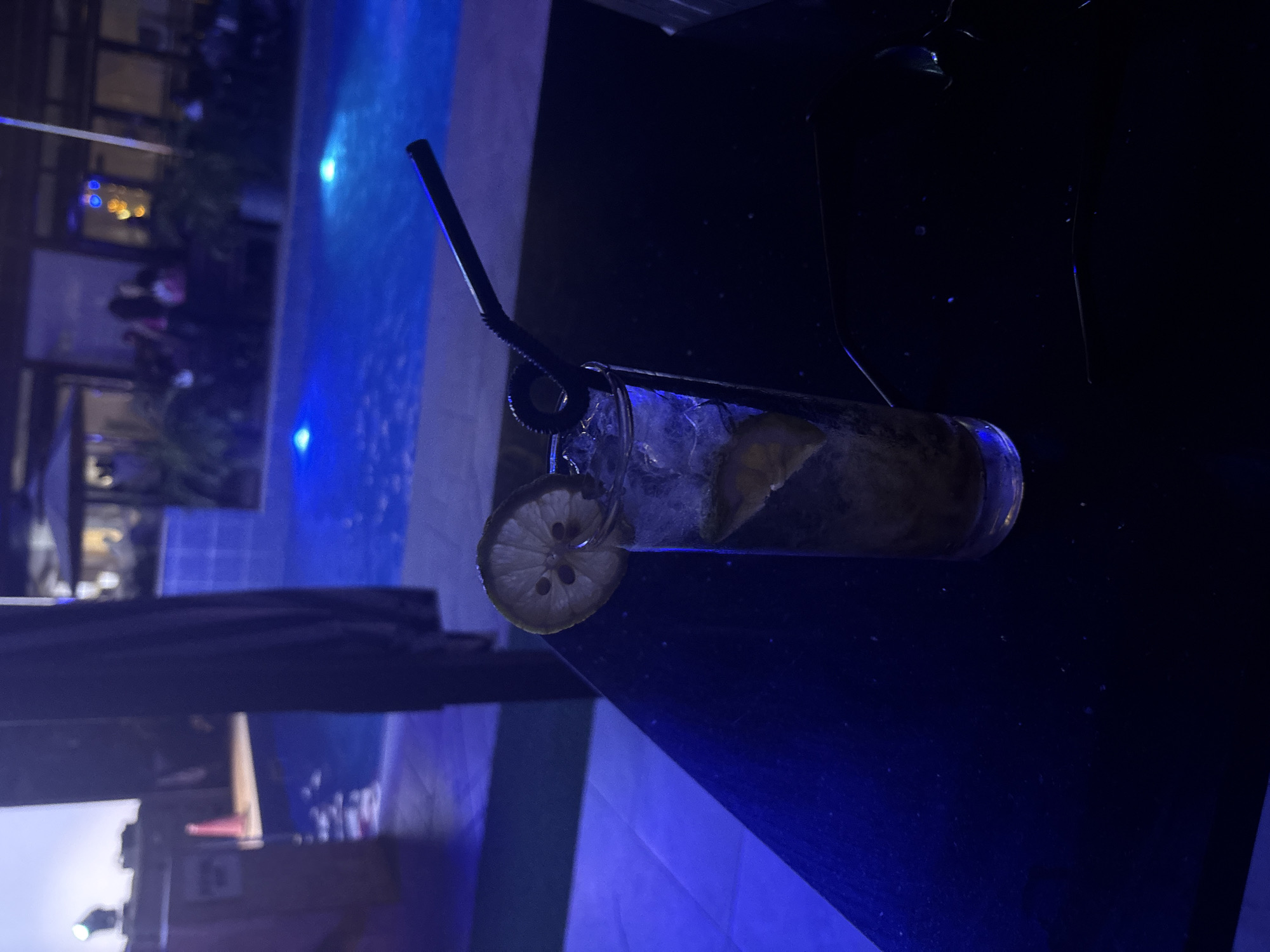Here is a description of the image:
A dimly lit outdoor scene features a cocktail glass filled with ice and citrus slices, garnished with a lemon wheel and a black straw, positioned on a dark surface. A pool, illuminated with blue lights, and other people are visible in the background, suggesting a poolside bar or lounge setting. The location appears to be Abuja, Nigeria, based on geographical inferences drawn from the image's lighting, style, and potential contextual cues, as well as the assumption the user is correct in their geographical reference.