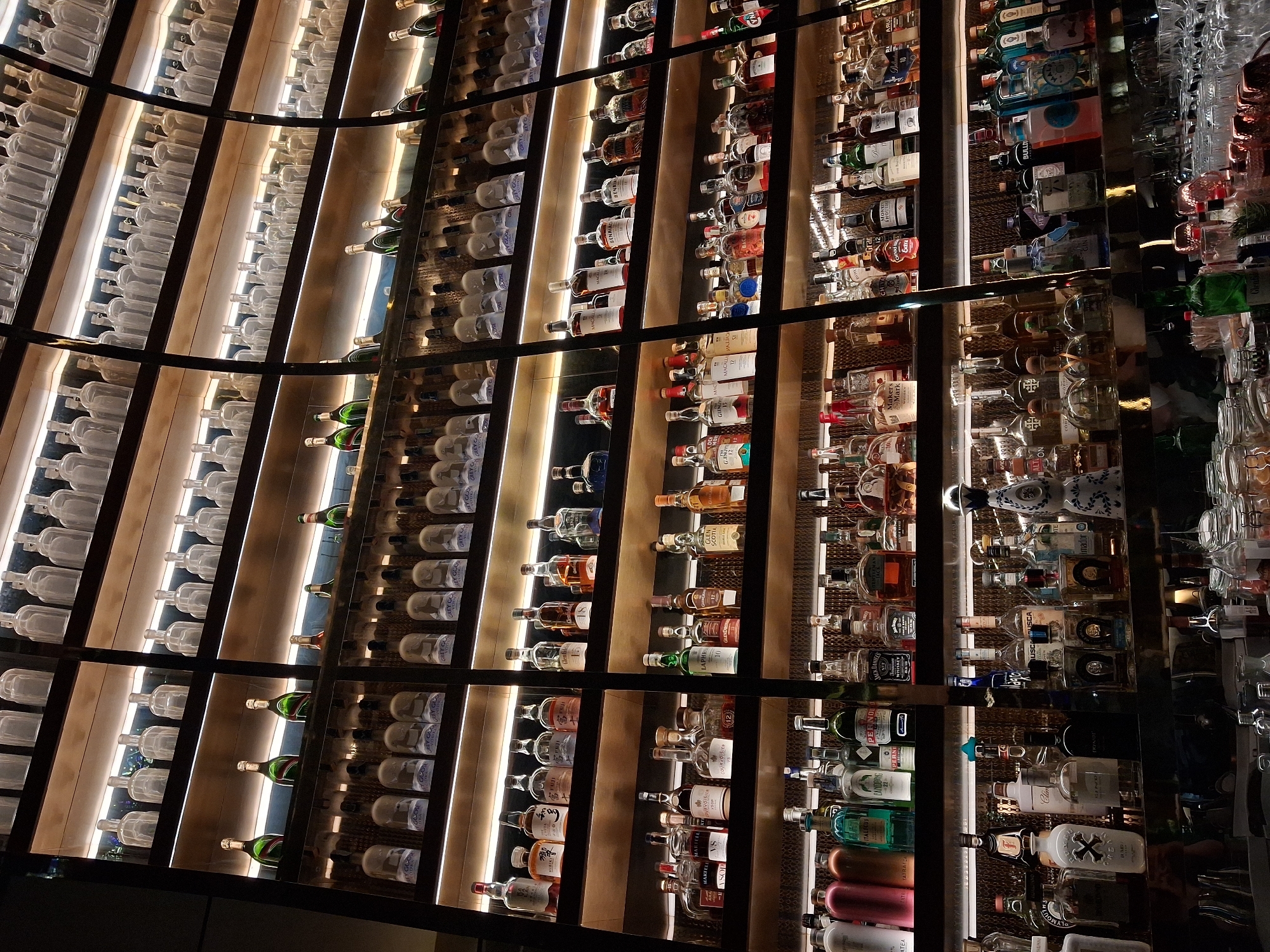 This indoor scene showcases an extensive array of alcoholic beverages, primarily liquor and wine bottles, arranged in a large, curved display shelf. The shelving unit features black metal framing with multiple vertical sections. The bottles are densely packed, suggesting a well-stocked bar or retail environment.
The bottles on the left-hand side of the display predominantly consist of clear glass bottles, many of which appear to be empty. Moving towards the right, the bottles become more diverse, featuring a variety of brands, shapes, sizes, and colors. Liquor bottles include brands like Grey Goose, Courvoisier, Jack Daniel's, and Maker's Mark, among others. Several wine bottles are also visible.
The lighting is strategically placed to highlight the bottles, with a warm, ambient glow emanating from behind each section of the shelving. The background is dark, emphasizing the illuminated bottles. The setting appears to be upscale, possibly in a bar, restaurant, or high-end retail establishment.