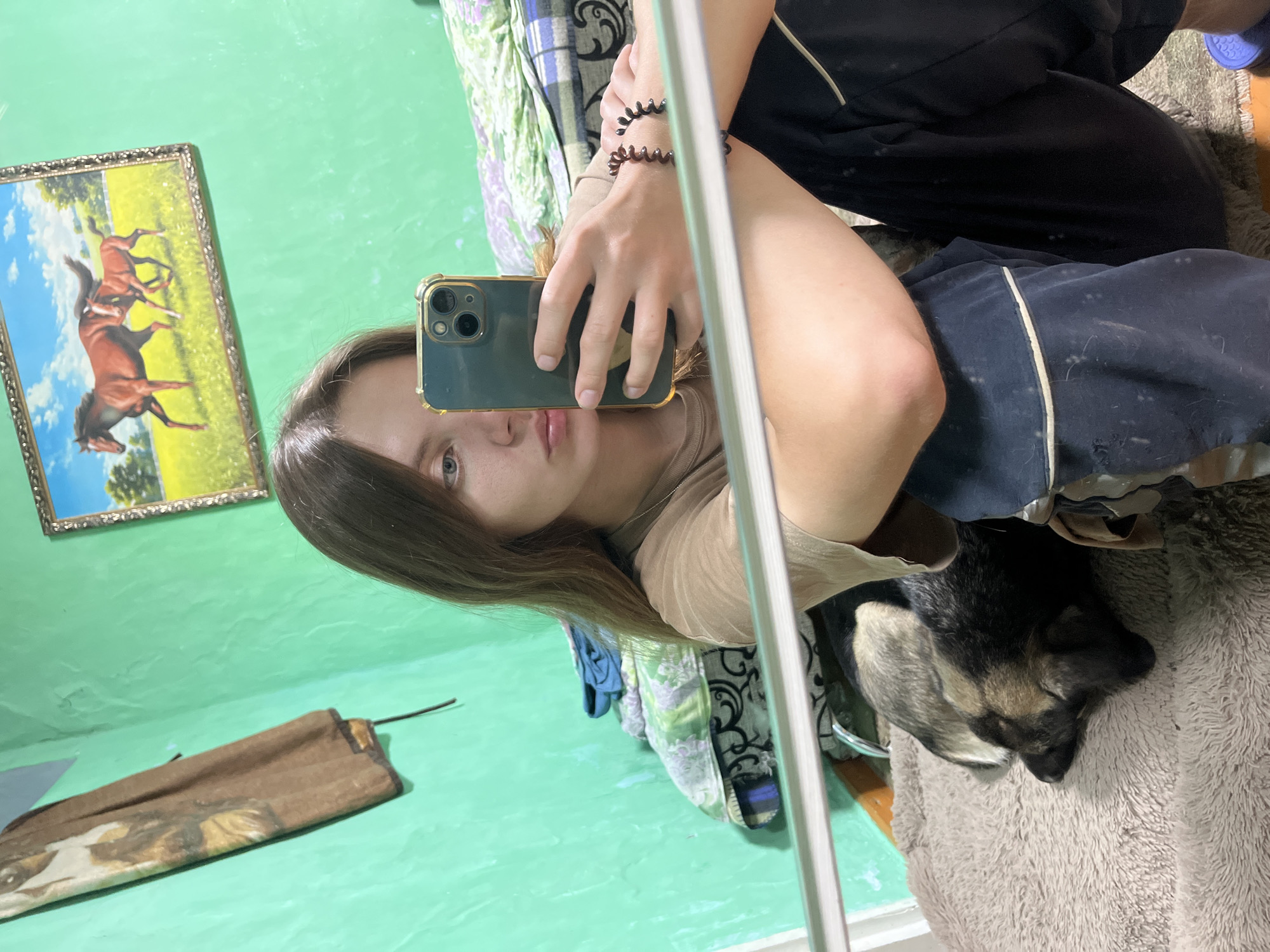 The image shows a young woman taking a selfie in what appears to be a bedroom. She is holding an iPhone and is wearing a light brown t-shirt and dark pants. To her left, a dog is sleeping on a fuzzy blanket. The room is painted a bright green and a framed picture of horses hangs on the wall. Other items visible include a pile of blankets and a hanging brown cloth. The scene is indoors and the lighting suggests it may be daytime or artificial lighting is in use. There is no information in the image to indicate the location is Nizhny Tagil, Russia.