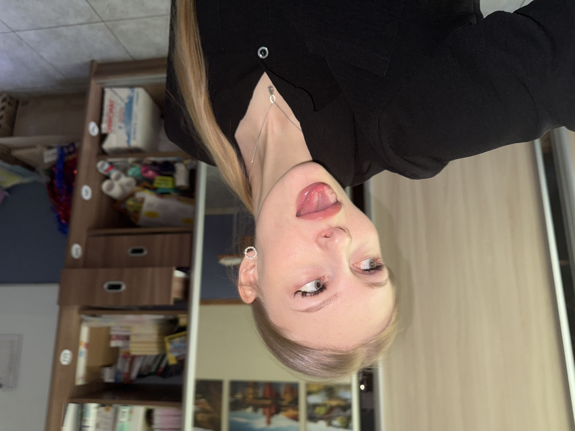 A young, fair-skinned woman with blonde hair is oriented upside down in the image frame. She has a pierced ear with a small, circular earring, and is wearing a silver necklace. Her mouth is slightly open, and her tongue is visible. She is wearing a black button-up shirt or jacket. Behind her, a shelving unit filled with books, boxes, and other objects is visible. A white door or panel with vertical lines is on the right side of the frame. Below the woman, there is what appears to be a series of art prints depicting landscape and figures. While I can note these objects in the image, I cannot infer a location such as Perm, Russia, as there is insufficient information for that determination.