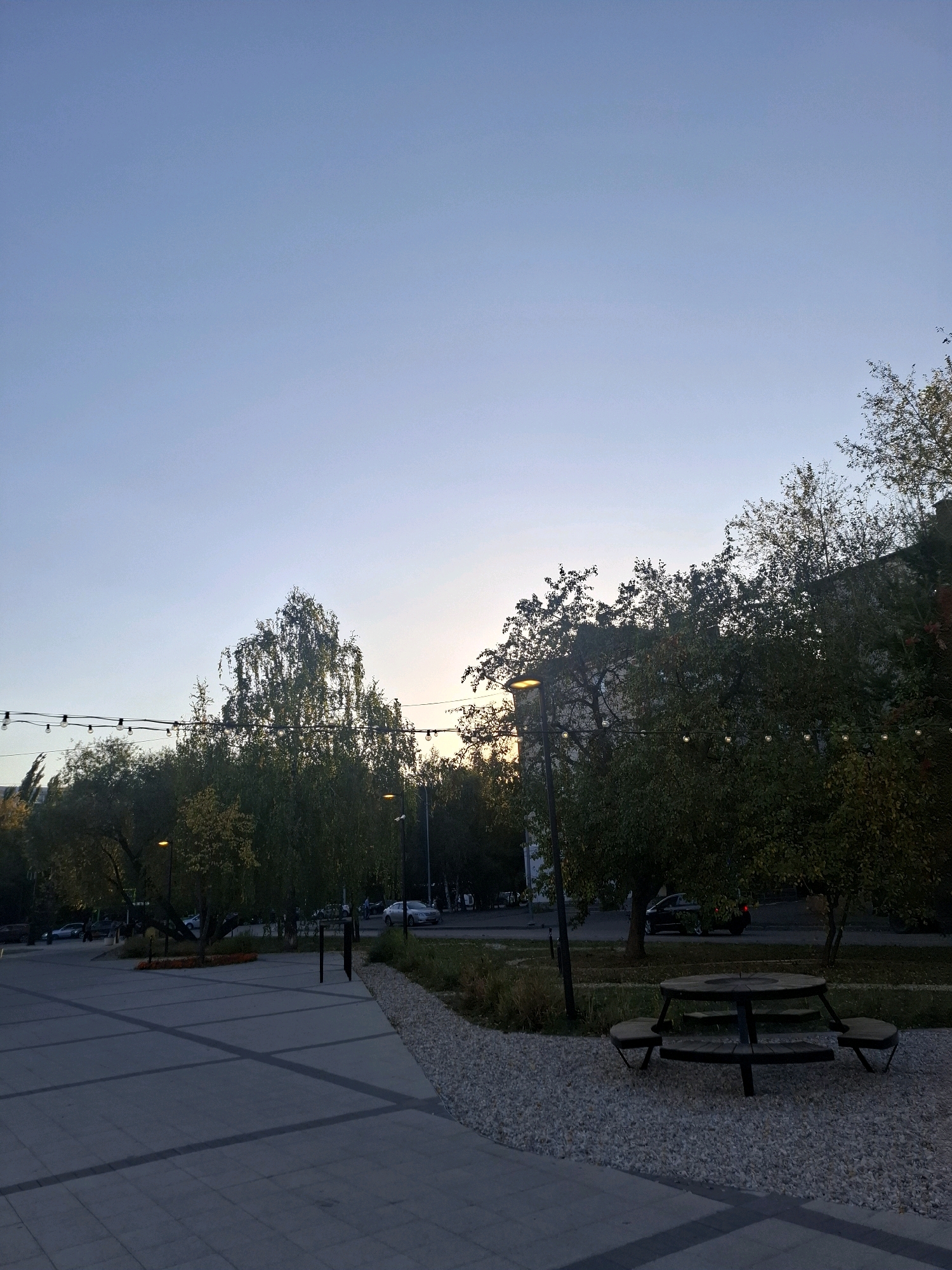 The image depicts an outdoor public space in the city of Tyumen, Russia, likely during twilight hours. The foreground is dominated by a large paved area with a geometric pattern of grey tiles. To the right, a gravel area features a wooden picnic table with attached benches. Beyond this, a line of trees with autumnal foliage separates the paved area from a street where several cars are visible, including a silver sedan and a dark SUV. Streetlights with warm illumination are present, as is a string of decorative lights suspended between poles across the trees. The background consists of buildings and more trees, all under a clear, pale blue sky transitioning from day to evening. There are no people visible in the image.