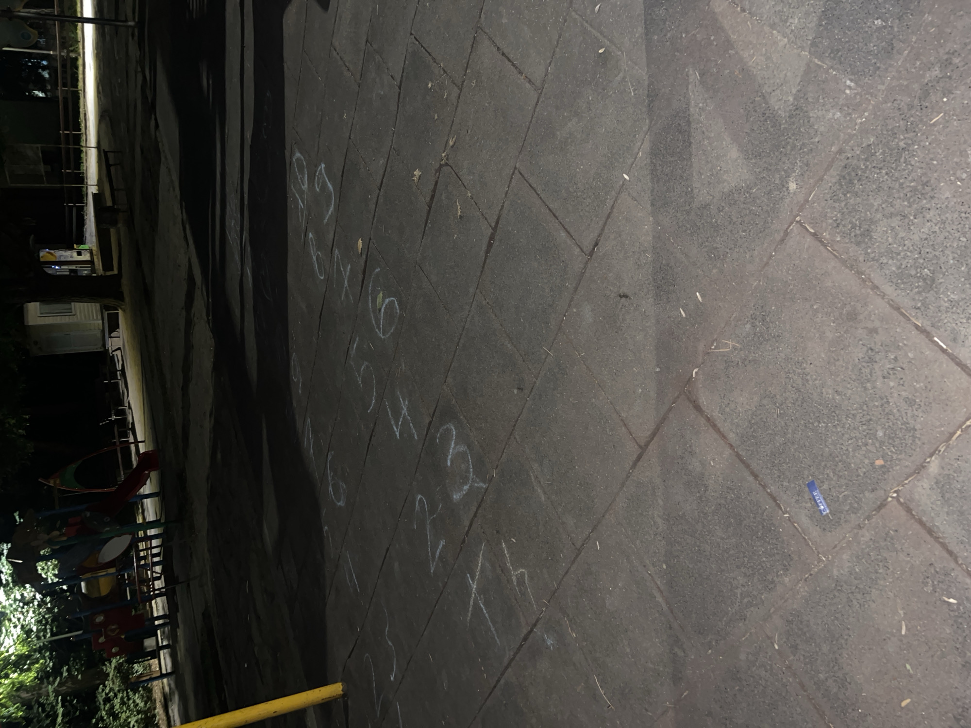 The image depicts a nighttime scene in a public park or playground area, likely in the city of Lovech, Bulgaria, given the context. The ground is paved with large, dark gray square tiles, and is illuminated by artificial lighting, casting long shadows.
Scattered across the pavement are several numbers drawn with white chalk, appearing to be part of a game or activity. The visible numbers include "3", "5", "6", "8", "9", "4", "2", and "1", suggesting a hopscotch-like pattern or sequence. There are also some faint circular chalk drawings.
In the background, a children's playground structure is partially visible, featuring colorful slides and climbing elements. Trees with dense foliage surround the area, with some branches and leaves illuminated by the lights. A yellow pole is positioned in the lower left corner of the frame.
In the middle ground, a small, light-colored structure, possibly a small building or kiosk, is visible, along with park benches. The overall impression is of a quiet, deserted public space at night. A small blue object, possibly a wrapper or piece of litter, is visible on the pavement in the lower center of the image.
