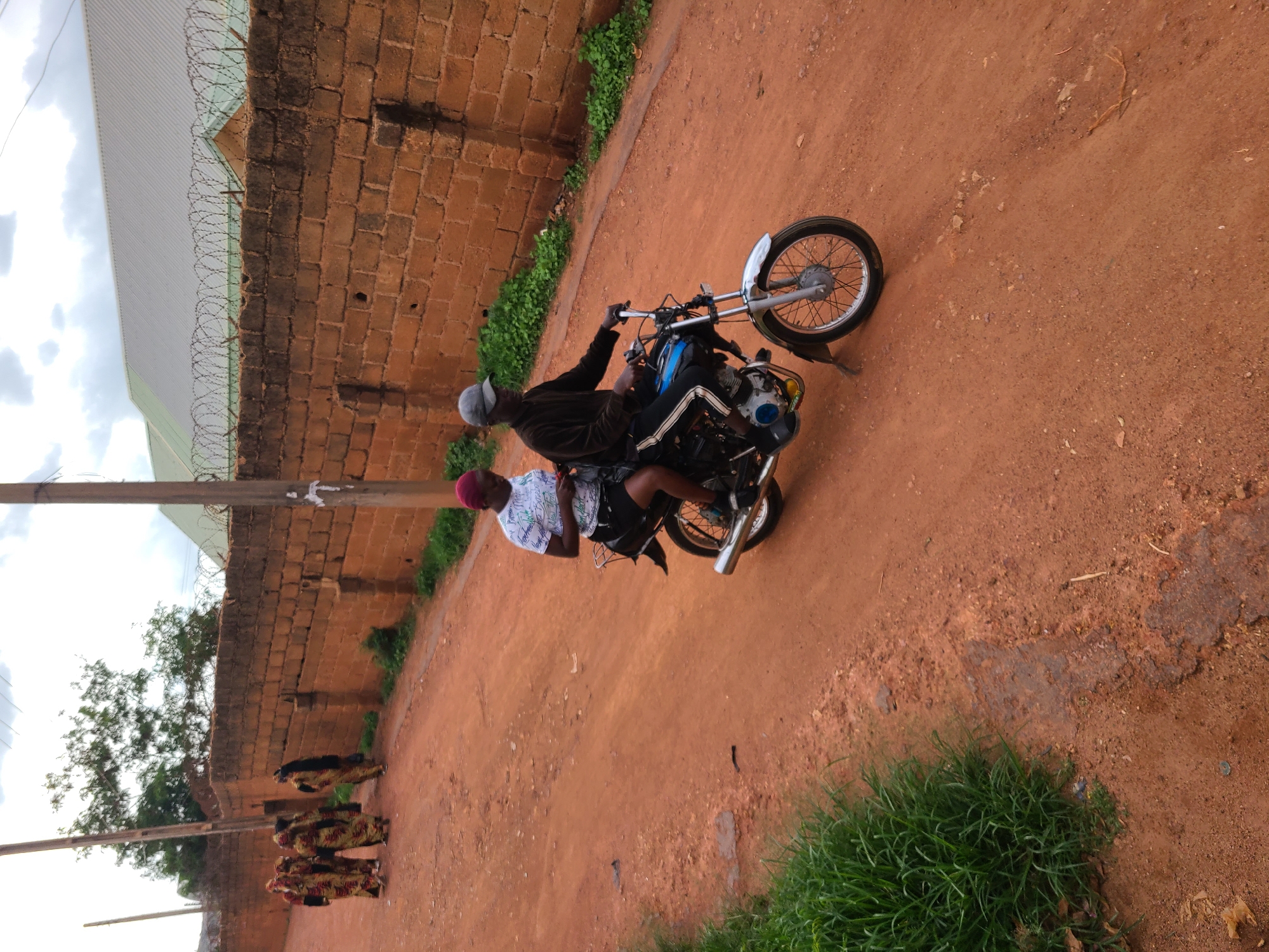 In Jos, Nigeria, a dirt road winds past a brick wall topped with barbed wire, leading to buildings with light-colored metal roofs. The road is predominantly reddish-brown soil, with patches of green grass growing along the left side. Two people are riding a motorcycle. The person in front, wearing a dark long-sleeved shirt and a grey cap, is driving. The passenger behind them, a person wearing a white t-shirt with text and a dark skirt, is looking down at something they are holding. In the background, on the left side of the image, a group of five people are walking along the road, all wearing colorful traditional clothing. A utility pole stands near the brick wall. The sky above is overcast.