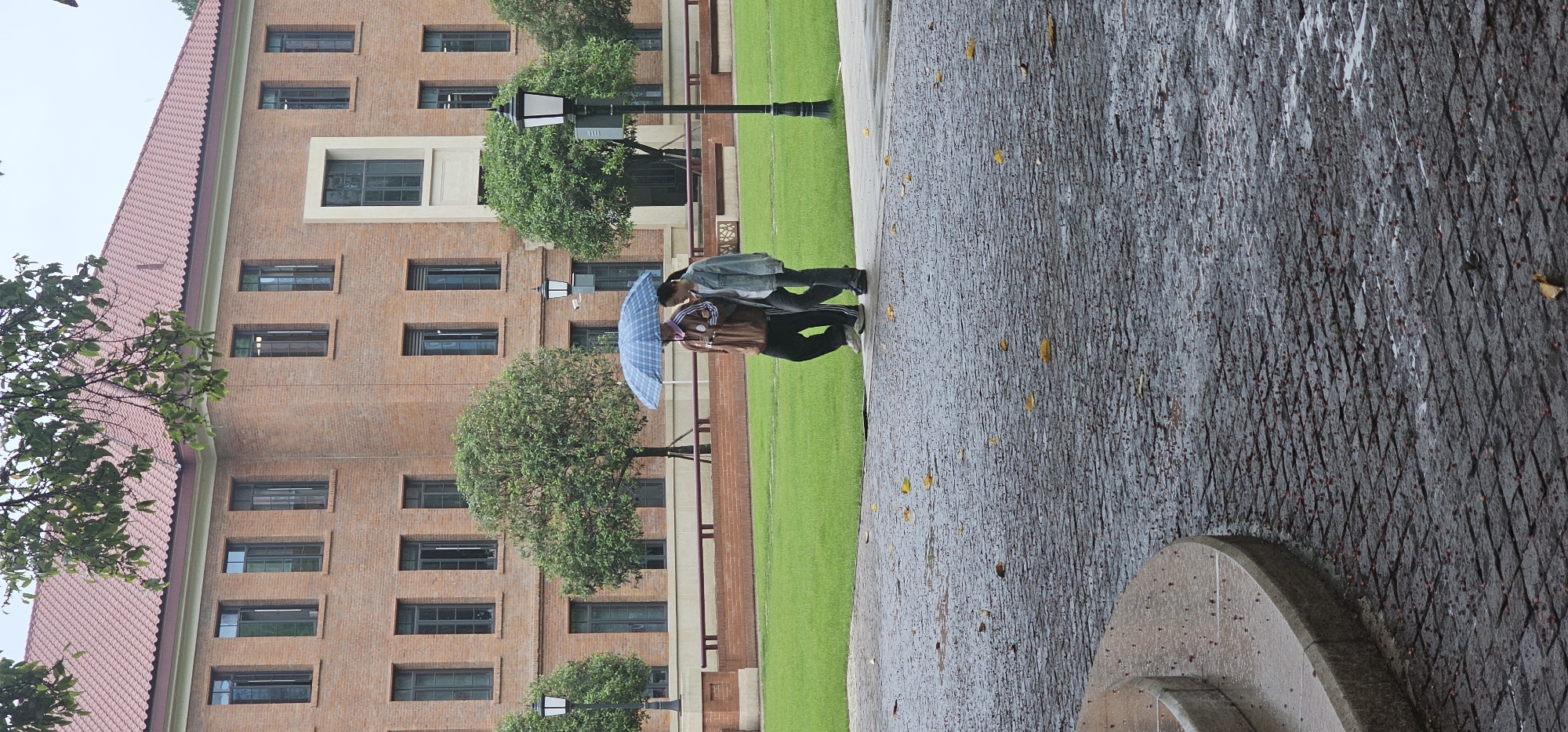 The image depicts two people under a blue umbrella on a rainy day. They are walking along a paved pathway adjacent to a green lawn. A large brick building with numerous windows and a red-tiled roof forms the backdrop. Street lamps are visible along the pathway. The scene is located in Yuzhong District, China. The pathway appears wet, indicative of rain.