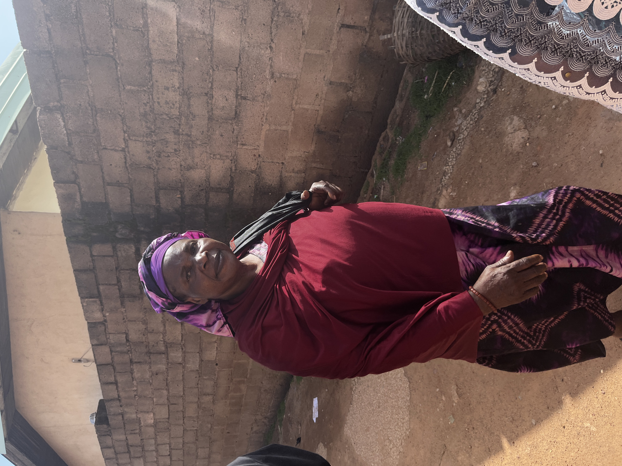 An older woman with a dark complexion stands outdoors, facing the camera. She is wearing a purple and pink headscarf, a deep red shawl draped over her shoulders, and a patterned skirt in shades of purple and black. A black bag is slung over her left shoulder. Her right hand, adorned with a reddish bracelet, rests on her lap, while her left hand holds the strap of the black bag. Behind her, a rough, grey brick wall forms the background, with a glimpse of a building roof and a pale wall above it. The ground is sandy and uneven, with sparse green vegetation and some scattered debris. A woven basket is visible on the right side of the frame. The scene appears to be in a modest urban environment, possibly a street or courtyard in Jos, Nigeria, given the architectural style and the woman's attire.