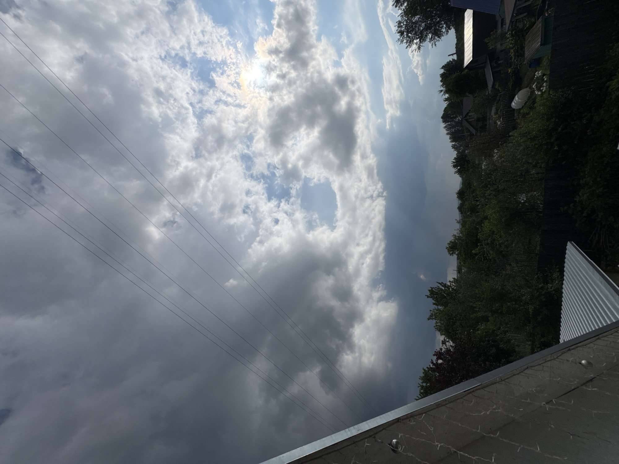 This is an outdoor, eye-level, vertical, medium shot photograph taken on a cloudy day, likely in Chaikovsky, Russia. The foreground features the corrugated metal roof and angled wall of a building, possibly a house or shed, with a reflective surface and visible rivets. In the mid-ground, a dense line of green trees, some with darker foliage, forms a natural barrier, interspersed with glimpses of houses and other structures, including what appears to be a greenhouse or protective covering. A dark wooden fence runs along the bottom of the mid-ground. The background is dominated by a dramatic sky filled with a mix of dark, heavy clouds on the left and brighter, scattered clouds with patches of blue sky and sunlight breaking through on the right. Several thin, dark power lines traverse the sky diagonally from the upper left to the upper right. No people are visible in the image.