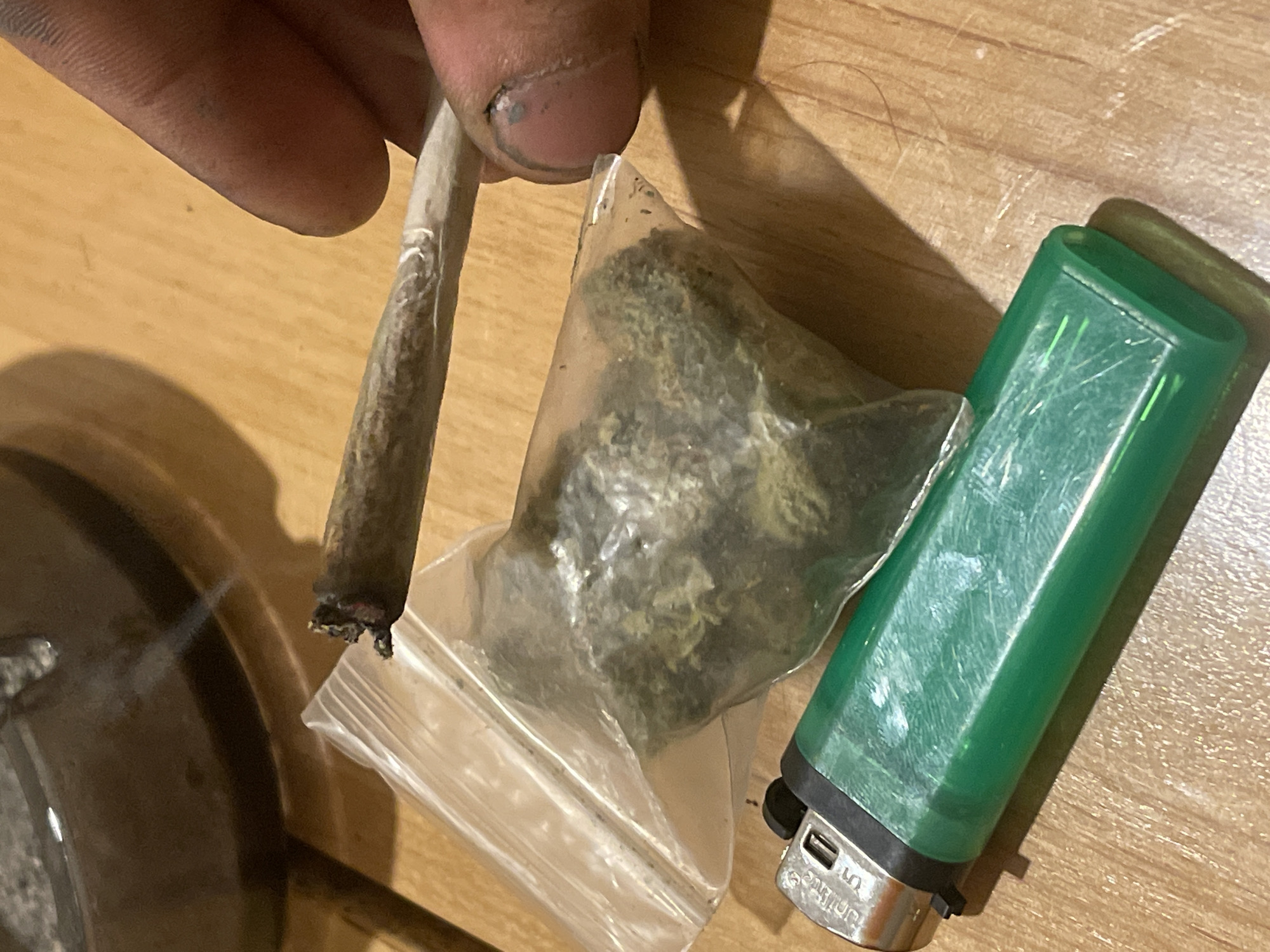 A hand is holding a lit marijuana cigarette over a plastic bag containing marijuana. A green lighter and an ashtray are also visible on a wooden surface. The scene is indoors, likely in a residential setting, with artificial lighting. The overall impression is one of casual drug use.