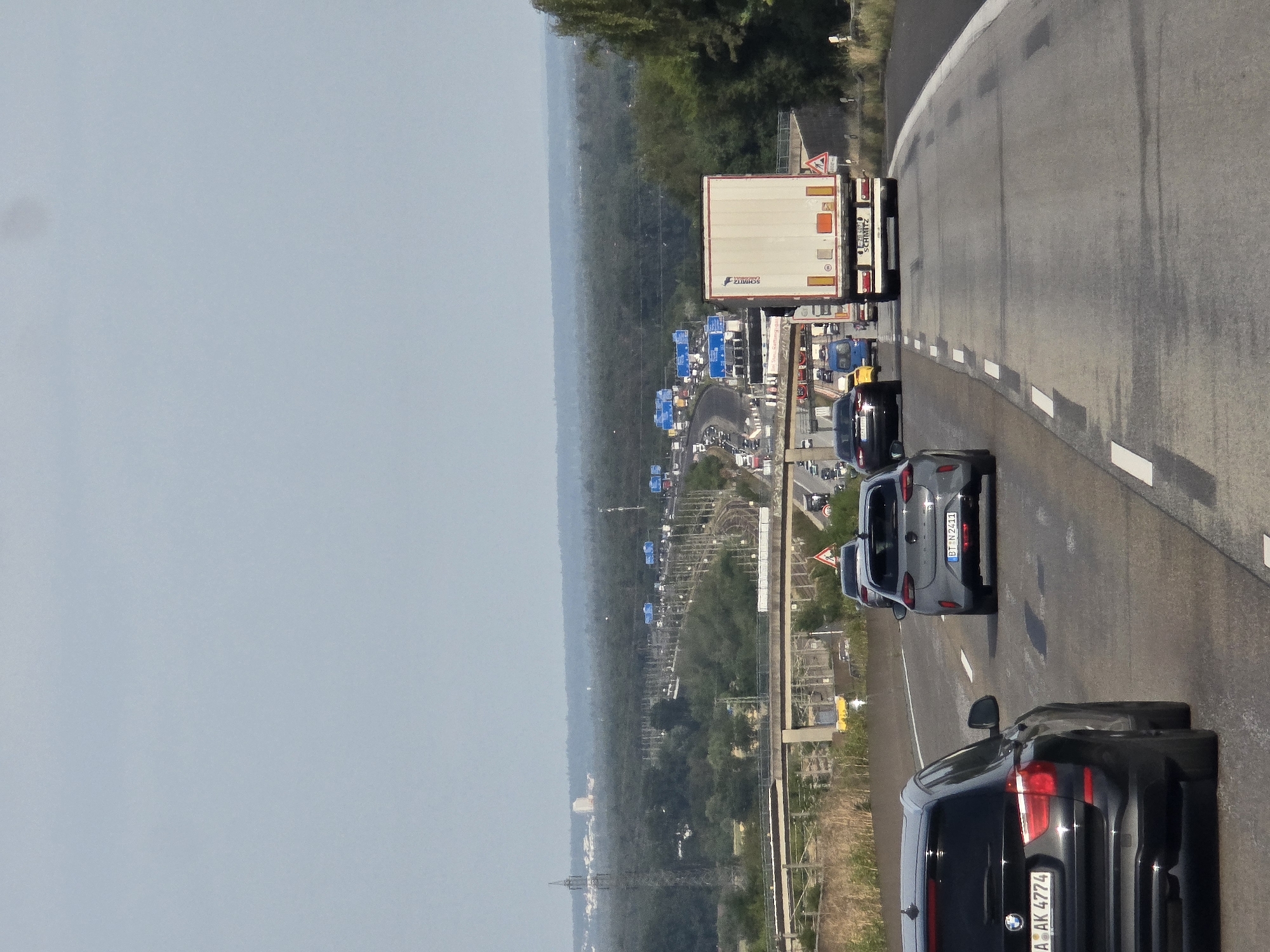 The image depicts a highway scene in Germany, with a focus on a traffic jam. Multiple vehicles, including cars and trucks, are visible on the asphalt road. The sky is overcast. In the distance, a dense forest borders the highway, with some industrial structures and power lines visible on the horizon. A concrete overpass crosses the highway further up ahead. The scene is set in Wicker, Germany.
