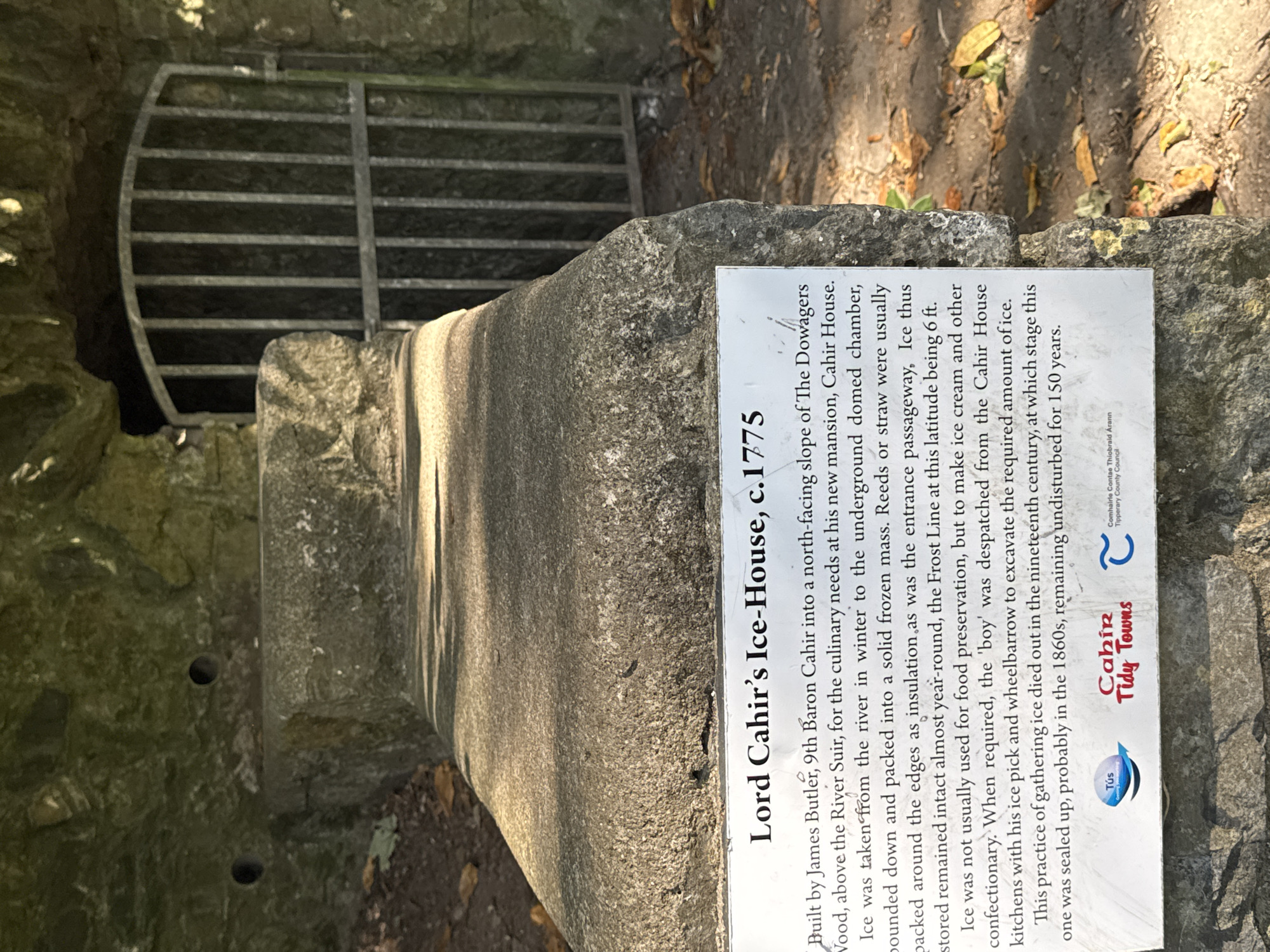 The image shows the Lord Cahir's Ice-House, located on the north-facing slope of The Dowagers Wood, above the River Suir, in Cahir, Ireland. The structure is built into the hillside and features a stone facade with an arched opening covered by a metal grate. A informational plaque in front of the ice-house details its history. The plaque states that it was built by James Butler, 9th Baron Cahir, around 1775, for the culinary needs of his new mansion, Cahir House. Ice was taken from the river in winter, packed into a solid frozen mass, and insulated with reeds or straw. The ice-house was designed to keep the ice stored year-round, with the Frost Line at this latitude being 6 ft deep. The practice of gathering ice ceased in the 19th century, and this particular ice-house was sealed up in the 1860s. The surrounding environment appears to be natural, with stone steps leading up to the ice-house and fallen leaves on the ground. The lighting suggests it is daytime, with sunlight casting shadows on the stone structure and the plaque.