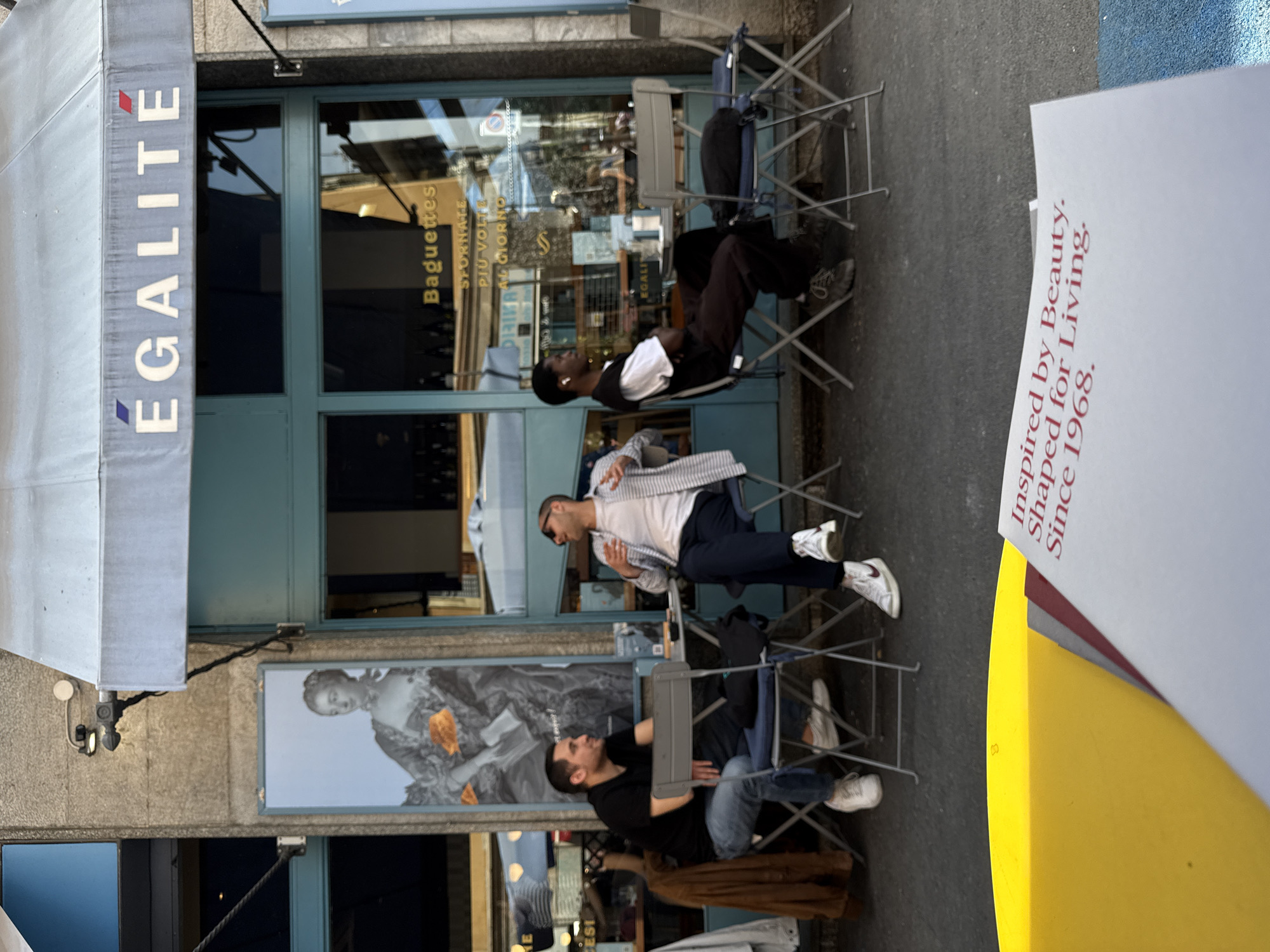 Three people are seated outside a building in what seems to be an outdoor cafe or restaurant. The storefront has a sign that says "Egalite" vertically on the left. The building signage indicates that "Baguettes" are sold and that they are "SFORNATE PIÙ VOLT AL GIORNO," also mentioning "EGALI." Two men are sitting at tables with chairs, facing each other and possibly conversing. A third person is seated at a table to the right of the other two. The environment is an urban setting, with the photograph seemingly taken during the day. The photo also has a sign saying "Inspired by Beauty. Shaped for Living. Since 1968". The cafe has a decorative image of a woman on the building. The city is Milan.