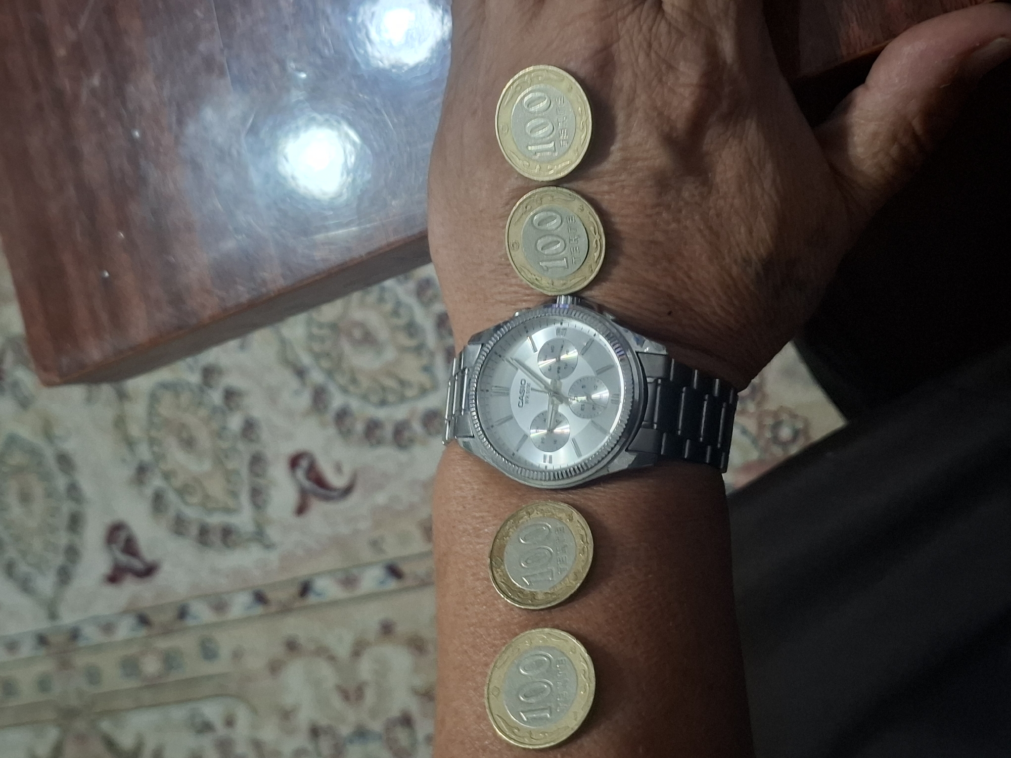 The image shows a close-up of a person's wrist and hand, adorned with a silver analog watch and four coins. The coins, all displaying the number "100" and text that appears to be "TENGE", are arranged in two pairs, with the watch positioned between them. The background suggests an indoor setting with a patterned carpet and a wooden surface, possibly a table. The lighting is somewhat diffused, creating reflections on the wooden surface. The overall scene is likely a personal snapshot rather than a location-specific scene in Almaty, Kazakhstan, as no distinctive landmarks or geographical features are visible.