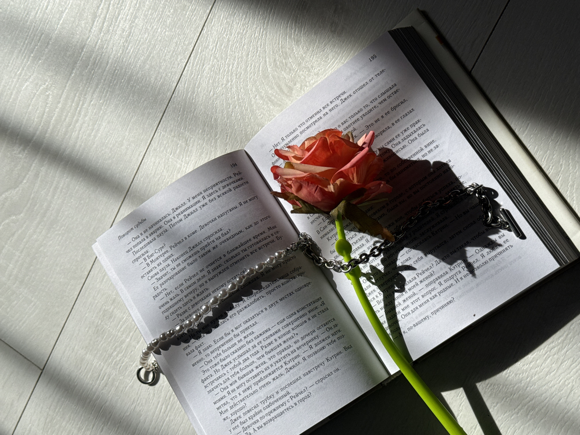 An open book with visible pages 194 and 195 lies diagonally on a light-colored surface, possibly wood or laminate, bathed in strong, directional sunlight. A red-orange rose with a green stem is placed across the spine of the book, its petals resting on the right page. A pearl bracelet and a silver chain bracelet are draped over the book, with the pearl bracelet near the left page and the silver chain extending towards the rose and the right page.
The scene appears to be set indoors, possibly near a window, given the distinct shadows cast by the book and the objects, suggesting bright morning or late afternoon light. The overall mood is calm and reflective, perhaps romantic, due to the presence of the rose and the act of reading.
The text in the book is in Russian. On page 194, the title "Поворот судьбы" (Turn of Fate) is visible. The content includes dialogue and narrative, with names such as Jill (Джилл), Rachel (Рейчел), Jack (Джек), and Catherine (Кэтрин) frequently appearing. Phrases like "в реанимации" (in intensive care), "В Биг-Суре?" (In Big Sur?), and "В Монтерее." (In Monterey.) are discernible, indicating a dramatic storyline involving an accident and medical emergency. The text discusses personal relationships, responsibilities, and emotional responses, such as "Ее разочарование было таким же очевидным, как до этого радость." (Her disappointment was as obvious as her joy before).