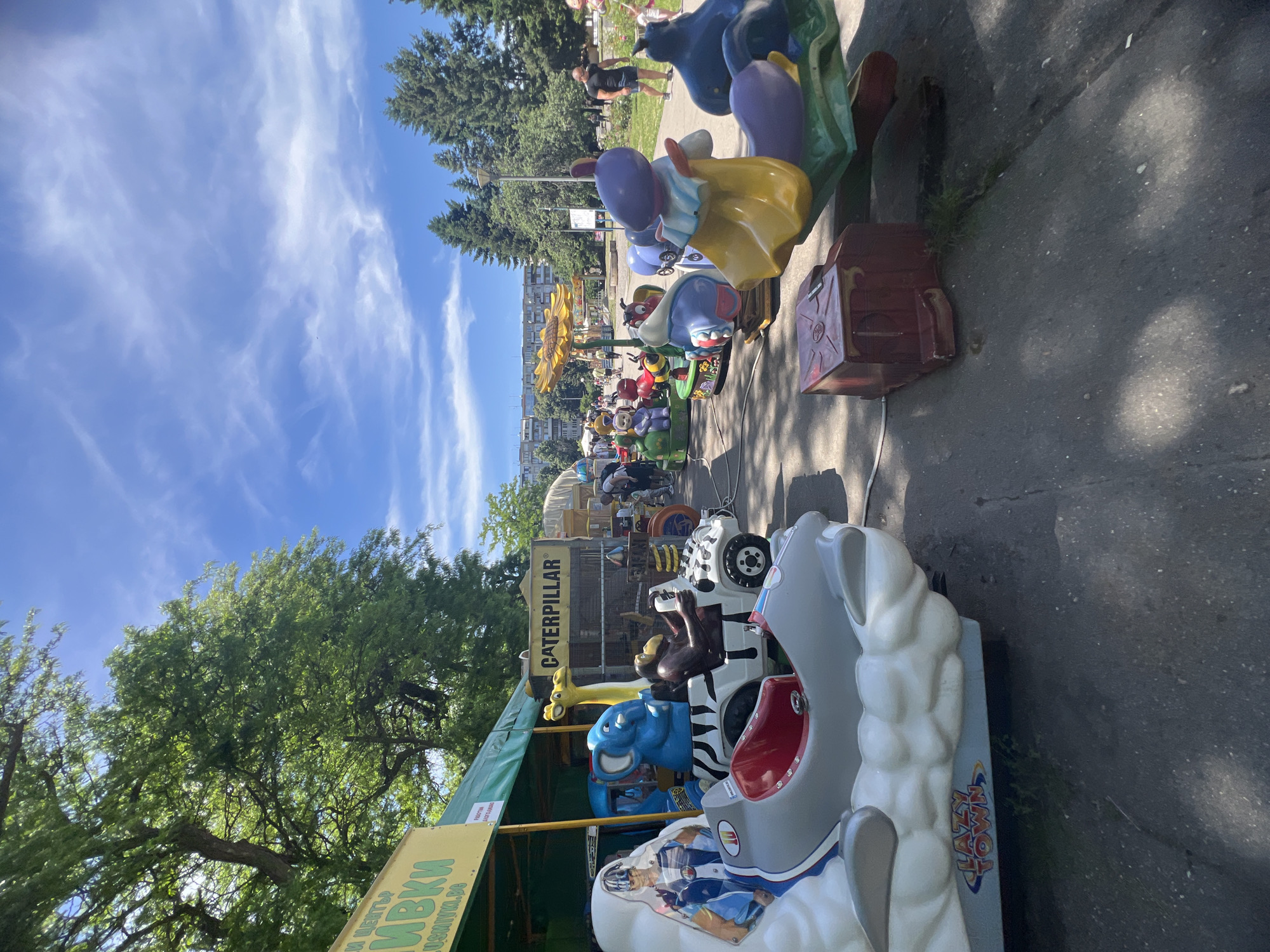 The image shows a row of kiddie rides located in a park in Sofia, Bulgaria. The rides depict various cartoon characters and animals, including a caterpillar with a sign reading "CATERPILLAR" above it. A vendor's stall is visible in the foreground. People can be seen walking in the background. The sky is blue with scattered clouds.