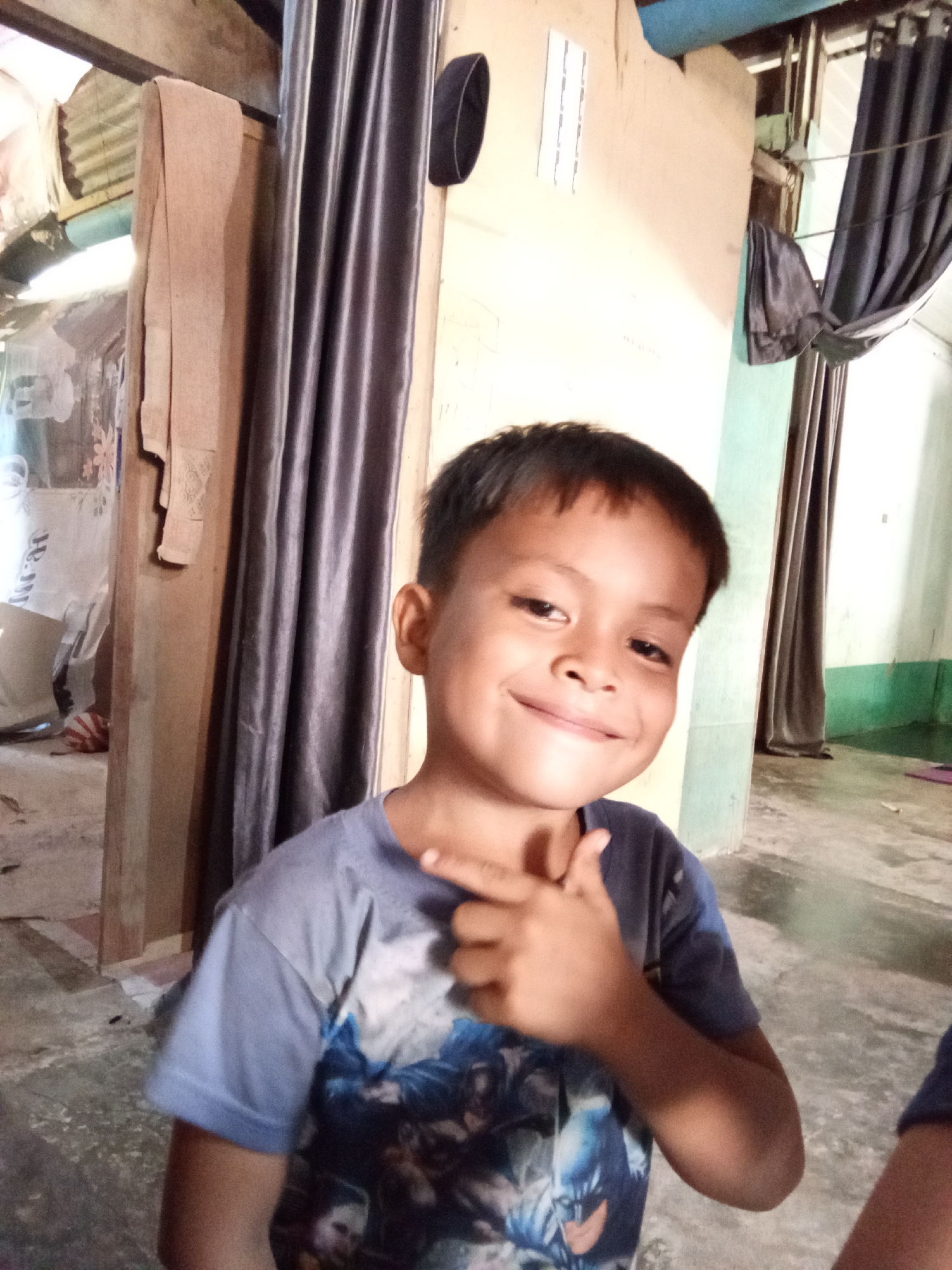 A young boy with short dark hair and a slight smile is in the foreground of this medium shot, positioned slightly to the left of the frame. He is wearing a short-sleeved, dark blue t-shirt with a graphic print on the front. His right arm is bent at the elbow, with his index finger extended and pointing upwards towards his face, as if he is about to say something. He is looking directly at the camera.
The background is dimly lit and appears to be an interior space. To the left of the boy, a tall, dark grey curtain hangs vertically, partially obscuring a wooden structure and some hanging fabric. A black cap is visible on top of the wooden structure. Further left, there is a glimpse of what appears to be laundry hanging in a room with corrugated metal walls. To the right of the boy, a light blue wall meets a light beige wall, with another dark grey curtain hanging in the background. The floor is concrete. The overall impression is a casual indoor setting.