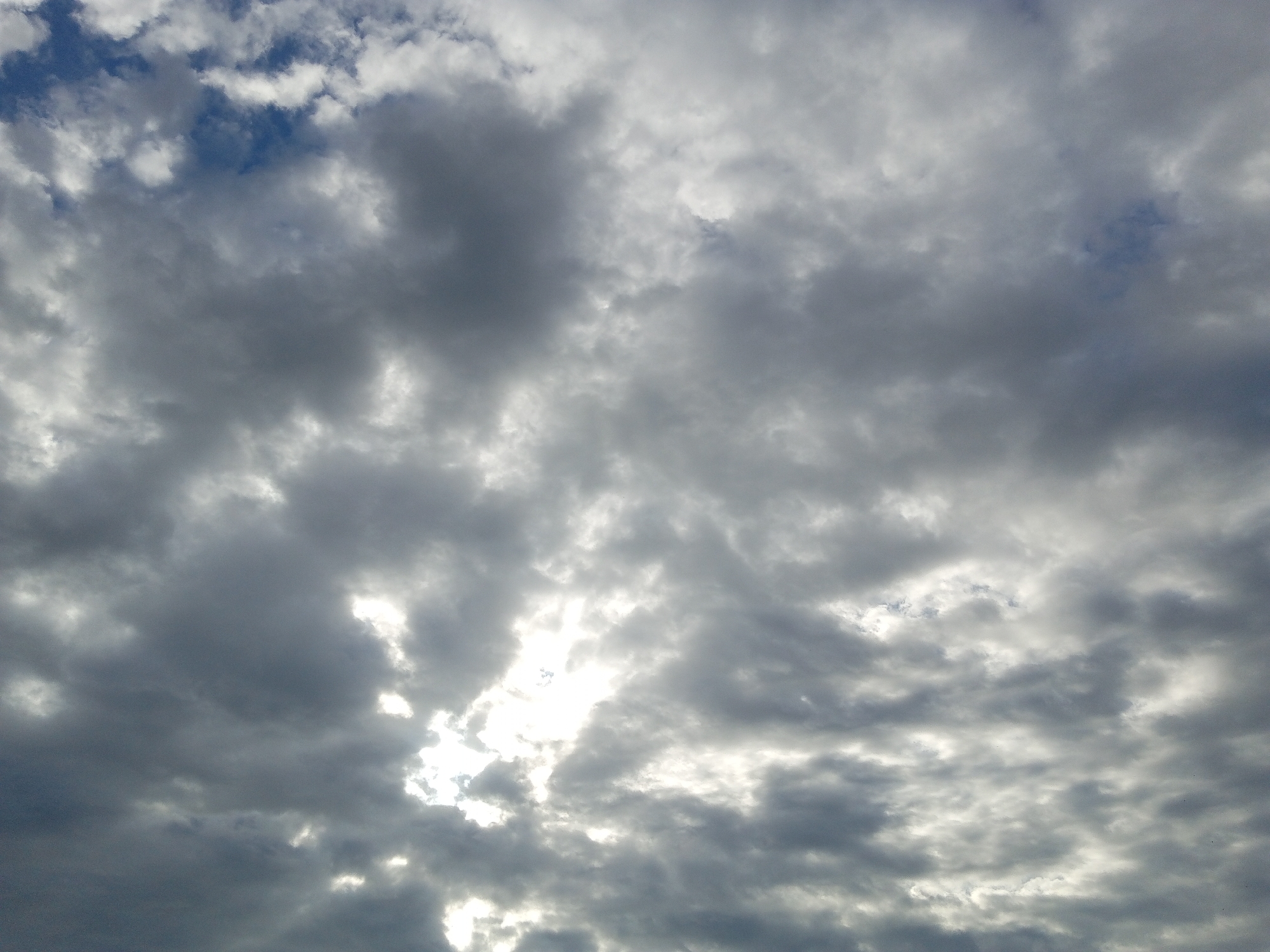 The image depicts an overcast sky. Dominating the view are layers of gray clouds of varying densities, with patches of brighter areas indicating the sun shining through the cloud cover. Small sections of blue sky are visible in the upper left corner. The scene provides no other information about Buketteukuh, Indonesia, or its environment. No people, subjects, objects, actions, or interactions are visible in the image.