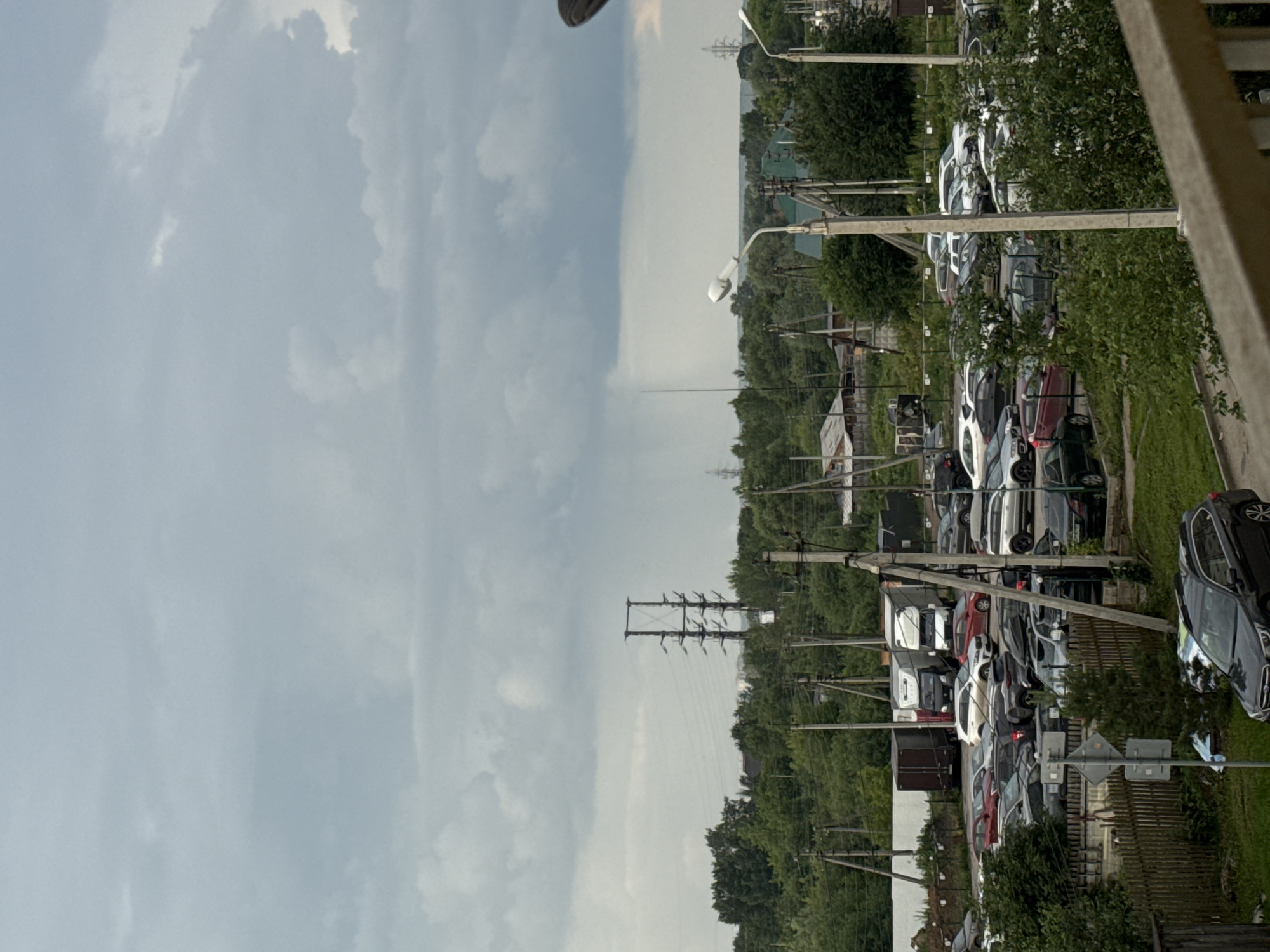 The image depicts an overcast sky over a suburban landscape that appears to be the village of Kondratovo, Russia. In the foreground, a wooden fence runs horizontally, and a dark SUV is parked on a grassy area. Beyond the fence is a parking lot filled with various vehicles, including cars, vans, and trucks. Several utility poles with wires are visible throughout the scene, supporting streetlights and electrical infrastructure. In the middle ground, a dense line of trees separates the parking lot from a more distant area with houses and more trees. In the background, more trees stretch to the horizon, and a power line tower stands prominently against the sky. A faint line of rain is visible in the distance under the clouds.