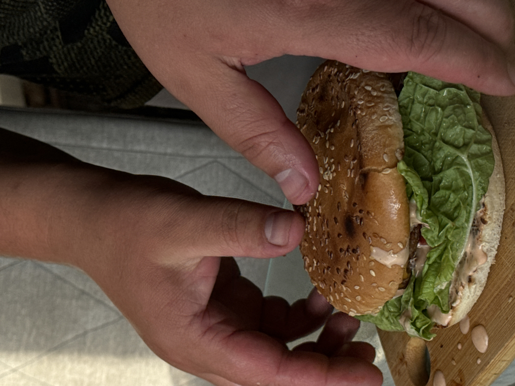 Two hands are shown in the foreground, holding a burger on a wooden cutting board. The burger has a sesame seed bun, lettuce, and sauce. The background is blurred and shows clothing. The image was taken in the city of Поназырево, Russia.