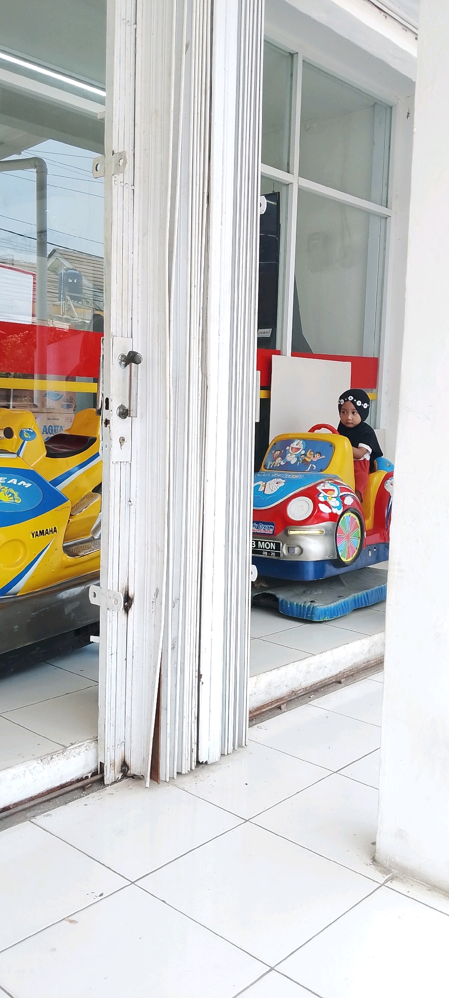 A child wearing a black hijab and black shirt is sitting in a small, multi-colored toy car with a cartoon character, Doraemon, depicted on its front. The car is parked on a blue platform on a tiled floor. To the left of the toy car, another yellow toy vehicle with "YAMAHA" written on it is partially visible. Both toy vehicles appear to be stationary amusement rides. The scene is framed by a white, accordion-style gate or door that is partially open, revealing the interior space. The background shows a glass storefront with reflections of buildings and the sky. The location is identified as Satriamekar, Indonesia.