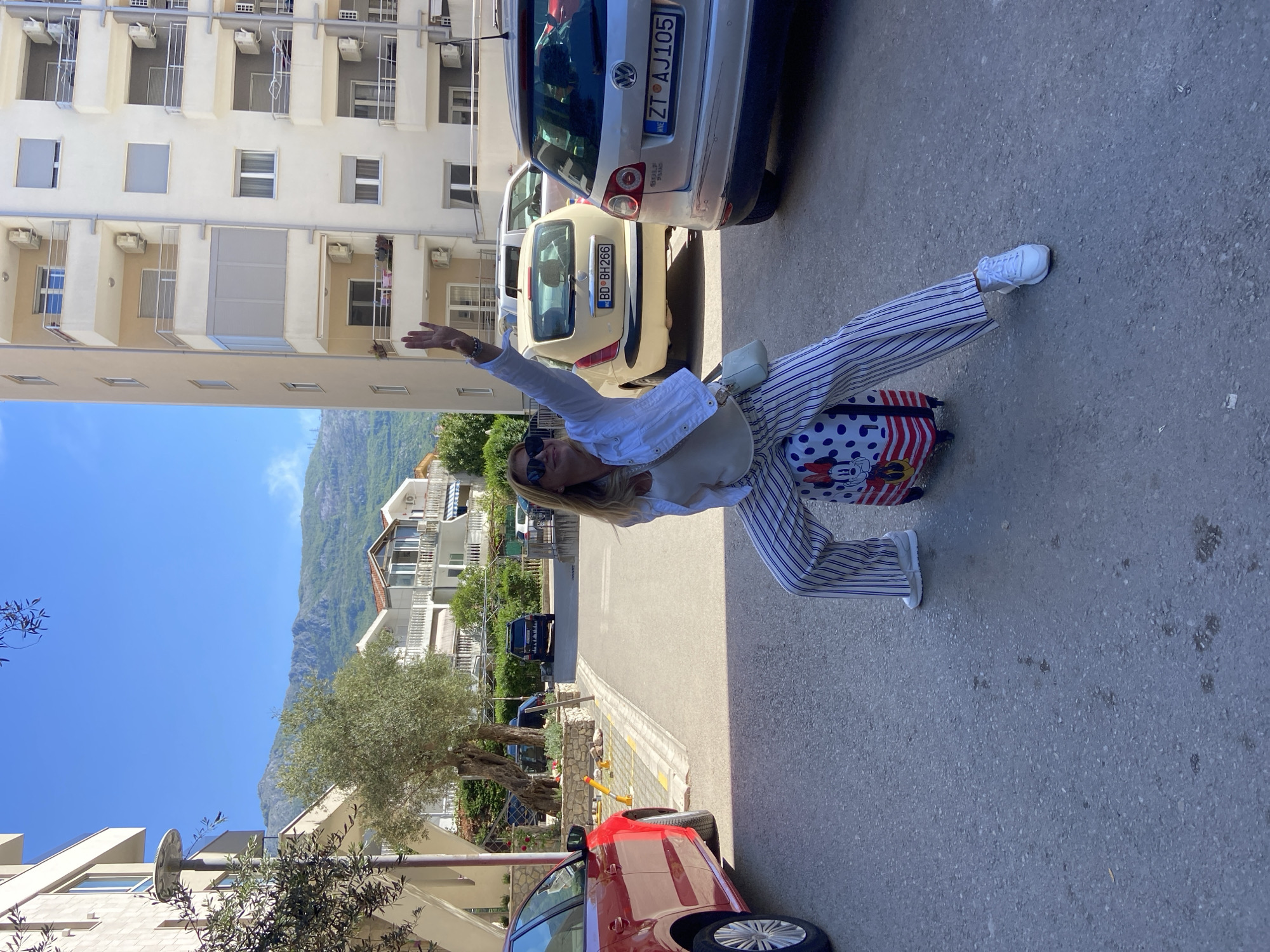 The image displays a bright, sunny street scene in Budva, Montenegro, likely during midday or early afternoon given the strong sunlight and distinct shadows. A blonde woman, wearing sunglasses, a white long-sleeved shirt, blue and white vertically striped pants, and white sneakers, is the central figure. She strikes a dynamic, playful pose, balancing on one foot with the other leg extended, and one arm raised as if waving or expressing joy. Behind her extended leg rests a small suitcase with a red, white, and blue polka-dot pattern, featuring Mickey Mouse and Minnie Mouse characters.
The setting is an urban street flanked by multi-story residential buildings, characterized by light-colored facades, balconies, and visible air conditioning units. The street is paved with asphalt. Several cars are parked along the roadside: a beige car with the Montenegrin license plate "BD BH266" (indicating Budva) is prominent, and behind it, a silver Volkswagen Golf with the Serbian license plate "ZT AJ105" is visible. The front wheel of a red car is seen in the immediate foreground.
Beyond the urban structures, lush green foliage and olive trees are scattered along the street. In the distant background, towering, rocky mountains rise under a clear blue sky, characteristic of the Montenegrin landscape. The scene evokes a sense of holiday or travel, with the woman's expressive pose suggesting happiness and relaxation.