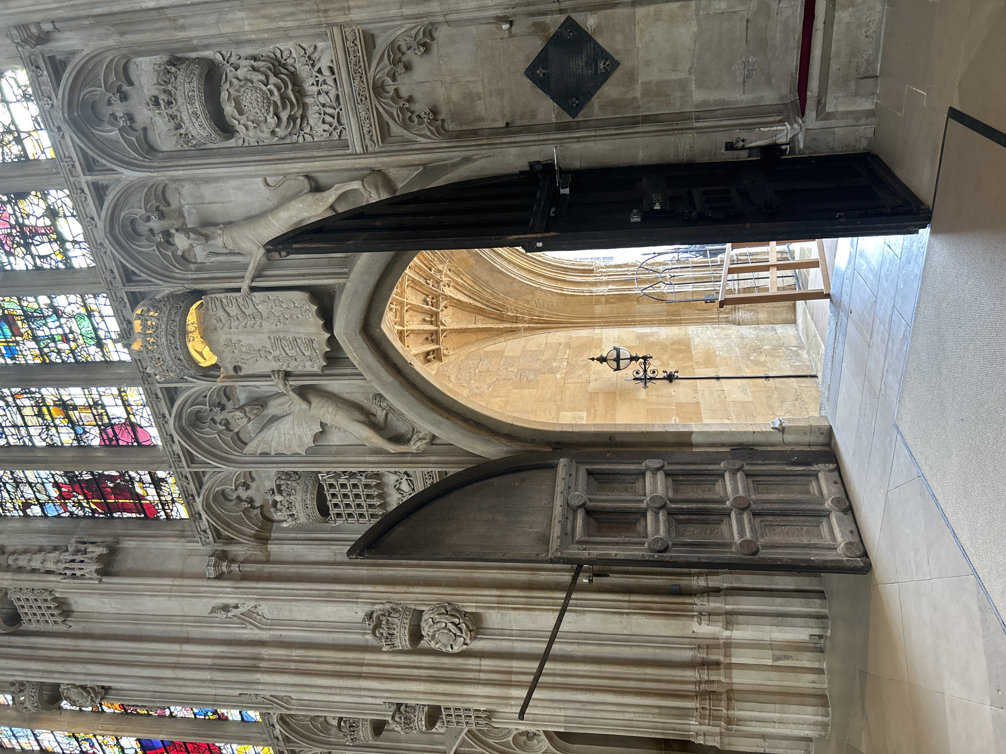 This image depicts an interior view of King's College Chapel in Cambridge, United Kingdom. The architecture features grand Gothic style with tall, fluted stone pillars and intricate carvings. Stained-glass windows with vibrant colors are visible at the top, letting in sunlight. In the foreground, two large, wooden doors are open, revealing an arched passageway beyond. The passageway has a stone vaulted ceiling and a lamp attached to a column. The floor is tiled with a textured rug placed near the entrance. Decorative elements on the stone walls include crowns, grilles, a rose motif, and carved mythical creatures. The overall impression is one of historical grandeur and intricate craftsmanship.