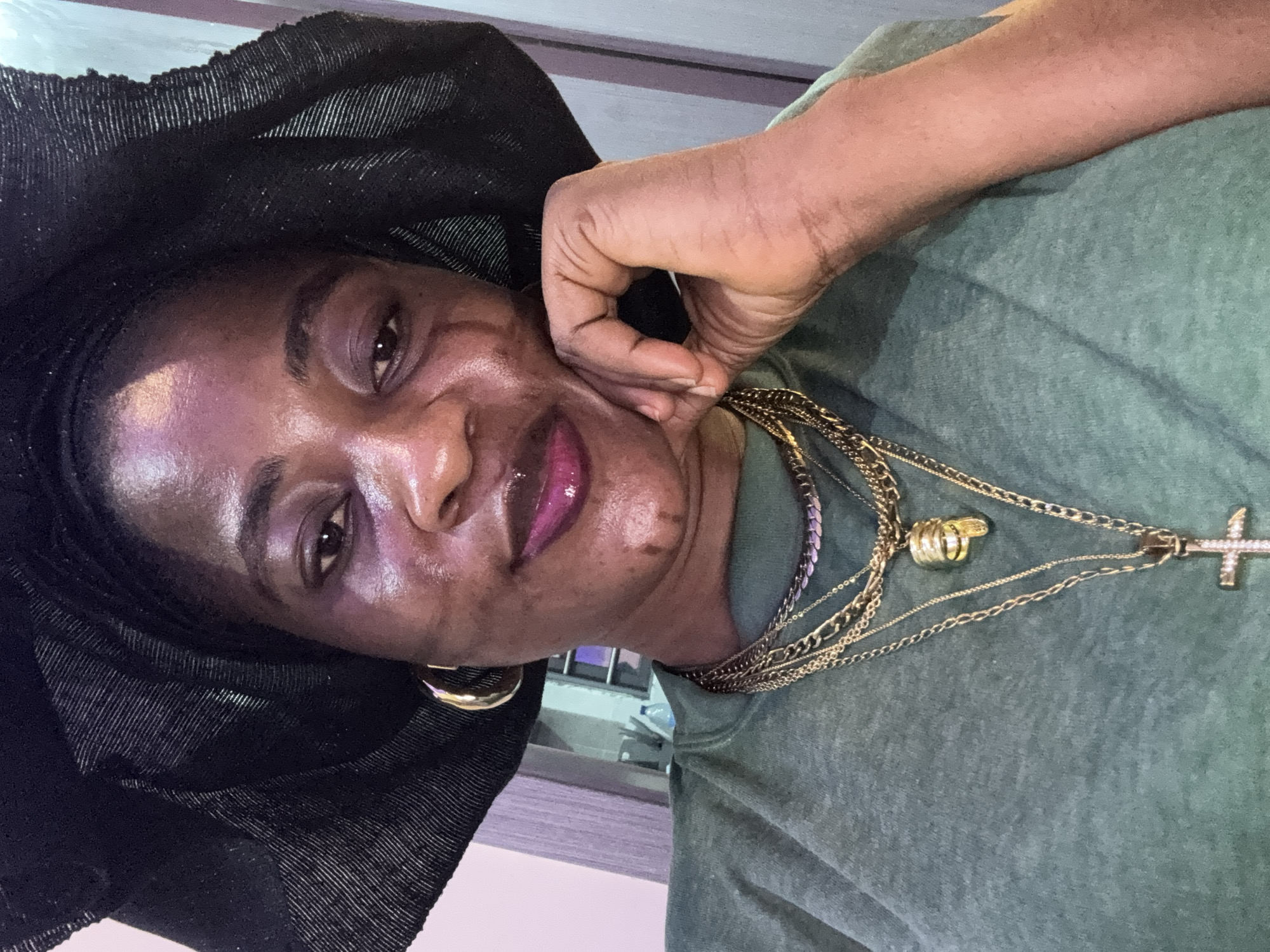 A portrait-style, close-up photograph shows a Black woman wearing a black headscarf and a green t-shirt. She is resting her chin on her hand, with her fingers gently touching her cheek. Her eyes are open and she appears to be looking towards the camera. She is wearing multiple layered gold necklaces, one of which has a crucifix pendant with clear gemstones. She also has a gold hoop earring in her visible ear. Her lips are colored with a glossy, bright pink lipstick. The lighting in the image is somewhat diffused, creating a subtle sheen on her skin. The background appears to be indoors, with a blurred hint of shelving or furniture. The overall mood of the image is intimate and perhaps contemplative.