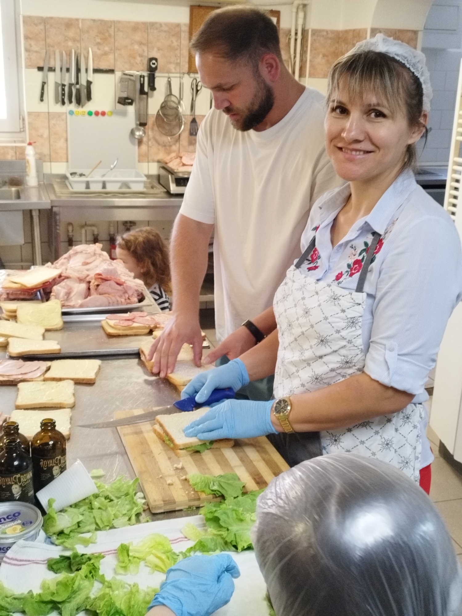 In a kitchen environment, potentially a catering facility or a large communal kitchen, two individuals are actively engaged in the preparation of sandwiches. The scene appears to be in Bucharest, Romania, based on the contextual information.
The foreground features a stainless steel countertop laden with ingredients for sandwiches. Slices of bread, some already spread with what appears to be butter or mayonnaise, are laid out. A pile of assorted meats, primarily ham and possibly other cold cuts, is visible on a tray in the background. Fresh lettuce leaves are scattered across the counter and on a white cloth. Two dark brown glass bottles, labeled "ROYAL CROWN COLA," and a small tub of what might be a spread or condiment are also present.
A woman, wearing a white hairnet and a light blue striped shirt with embroidered flowers, is smiling at the camera. She has an apron over her shirt and is wearing light blue gloves. She is in the process of assembling a sandwich, with her gloved hands holding a sandwich and a knife.
Beside her, a man with a beard, wearing a white t-shirt, is also working on sandwiches. He is also wearing light blue gloves and is seen with his hands on a sandwich. His focus is on the task at hand.
In the background, a young child with brown hair is partially visible, looking towards the meat. The kitchen walls are tiled with beige and brown square tiles. Various kitchen utensils, including knives hanging on a magnetic strip, ladles, and strainers, are mounted on the wall or resting on shelves. A stainless steel sink and other kitchen equipment are also discernible. The overall impression is one of efficient food preparation in a functional kitchen setting.