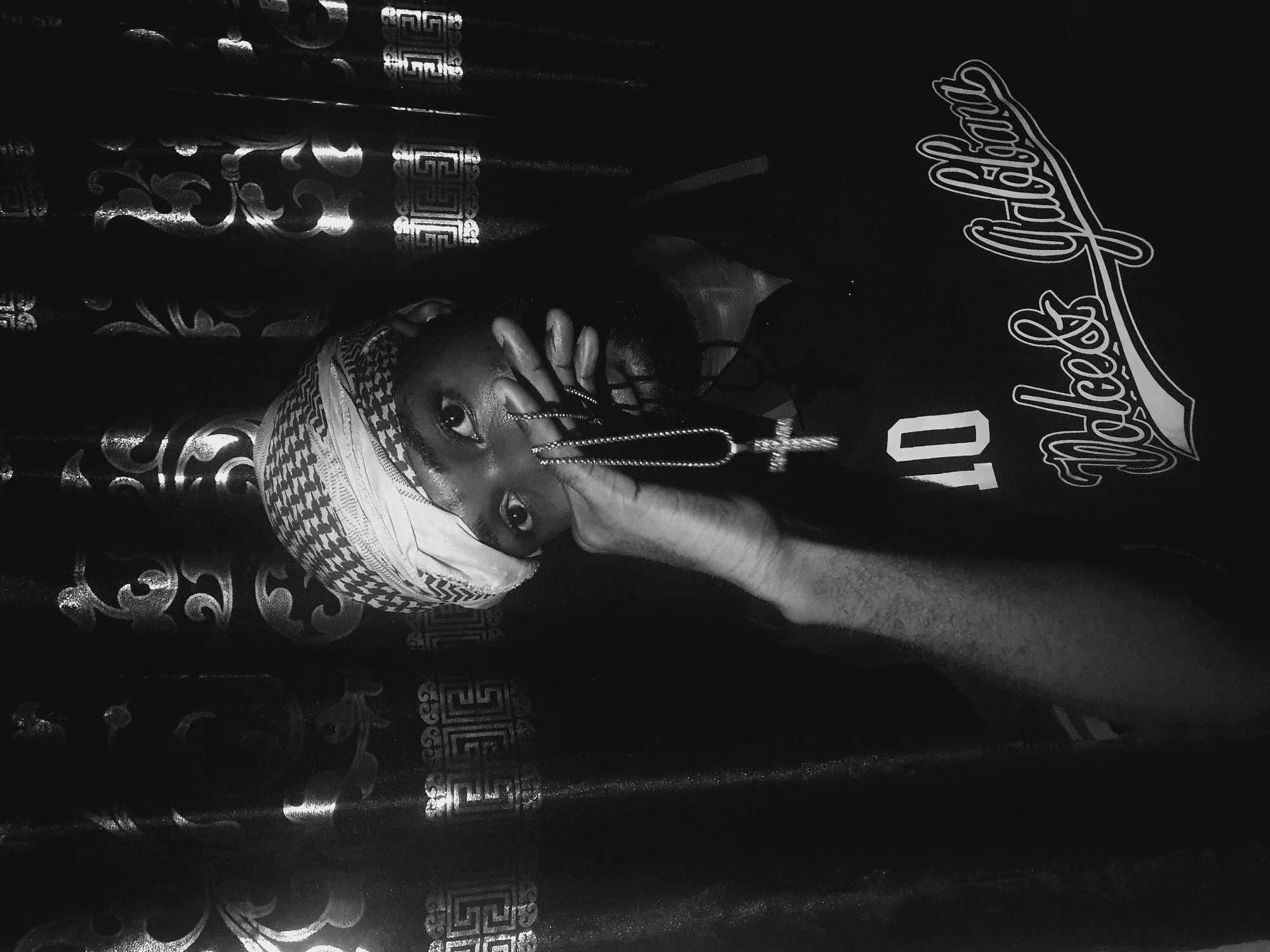 A black and white, horizontally oriented image shows a person wearing a patterned head covering partially obscuring their face, and a dark t-shirt with white lettering that reads "Dolce & Gabbana" with a number "10" beneath. They are holding a silver chain with a cross pendant near their face. The person appears to be lying on a surface with a repeating, ornate pattern. There is no discernible information in the image indicating that the location is Lokoja, Nigeria.