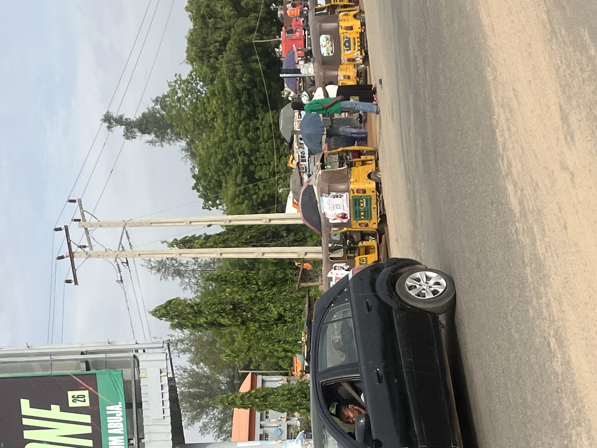 The image shows a street scene in Galadimawa, Nigeria. On the left side, there is a black car and a sign that reads "26" and "UM ABUJA". A person is visible inside the car. Along the roadside, there are numerous yellow auto-rickshaws parked. Several people are standing or walking near the auto-rickshaws. The background includes trees and power lines against a cloudy sky. The road surface is light-colored.