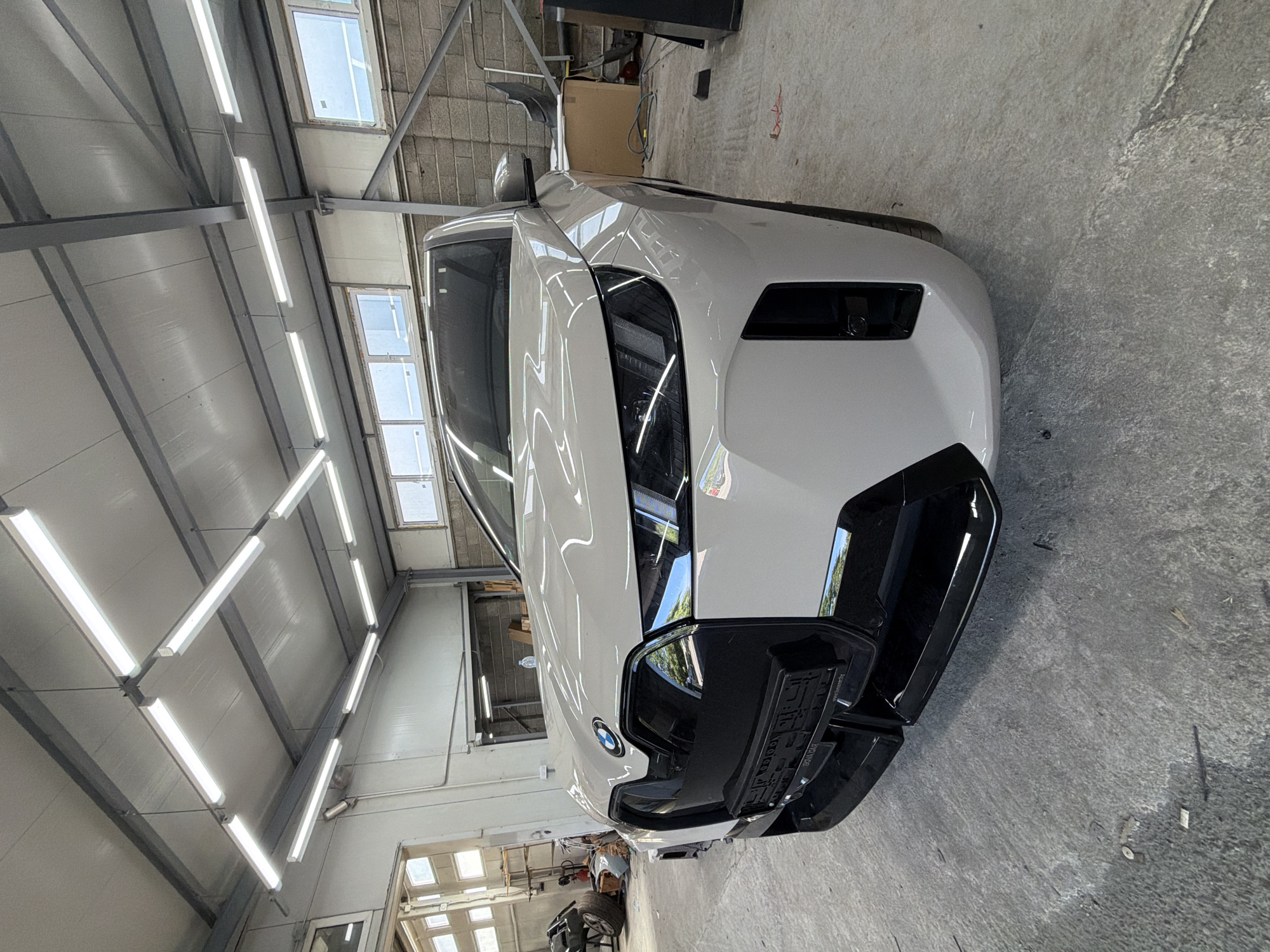 The image shows a white BMW car in a workshop or garage. The car is the primary subject, with its front end prominently displayed. It appears to be a newer model, possibly an electric vehicle, given its sleek design and large, dark grille. The car has a black accent on its bumper and a license plate holder attached.
The setting is an indoor workshop with a concrete floor and a metal-framed ceiling. The ceiling is lined with white panels and equipped with multiple fluorescent tube lights, indicating it's likely a workspace. Natural light is also entering from windows in the background, which appear to be small and rectangular. A spare tire and some tools are visible in the lower left corner of the image, further suggesting a repair or detailing environment. The overall lighting is bright and even, typical of a professional workshop.
There are no people visible in the image. The activity taking place is implied to be related to the car, such as maintenance, repair, or detailing. The car is stationary, and the focus is on its appearance and the environment it's in. The time of day cannot be determined from the image, but the presence of artificial lighting suggests it could be any time. The weather is not discernible as the scene is indoors.
A small BMW logo is visible on the front of the car. There's also text on the license plate holder, but it's not legible.
The location context of Burgas, Bulgaria, is provided, but there are no specific visual cues in the image that directly confirm this location. The workshop appears generic and could be found in many industrial or commercial areas.