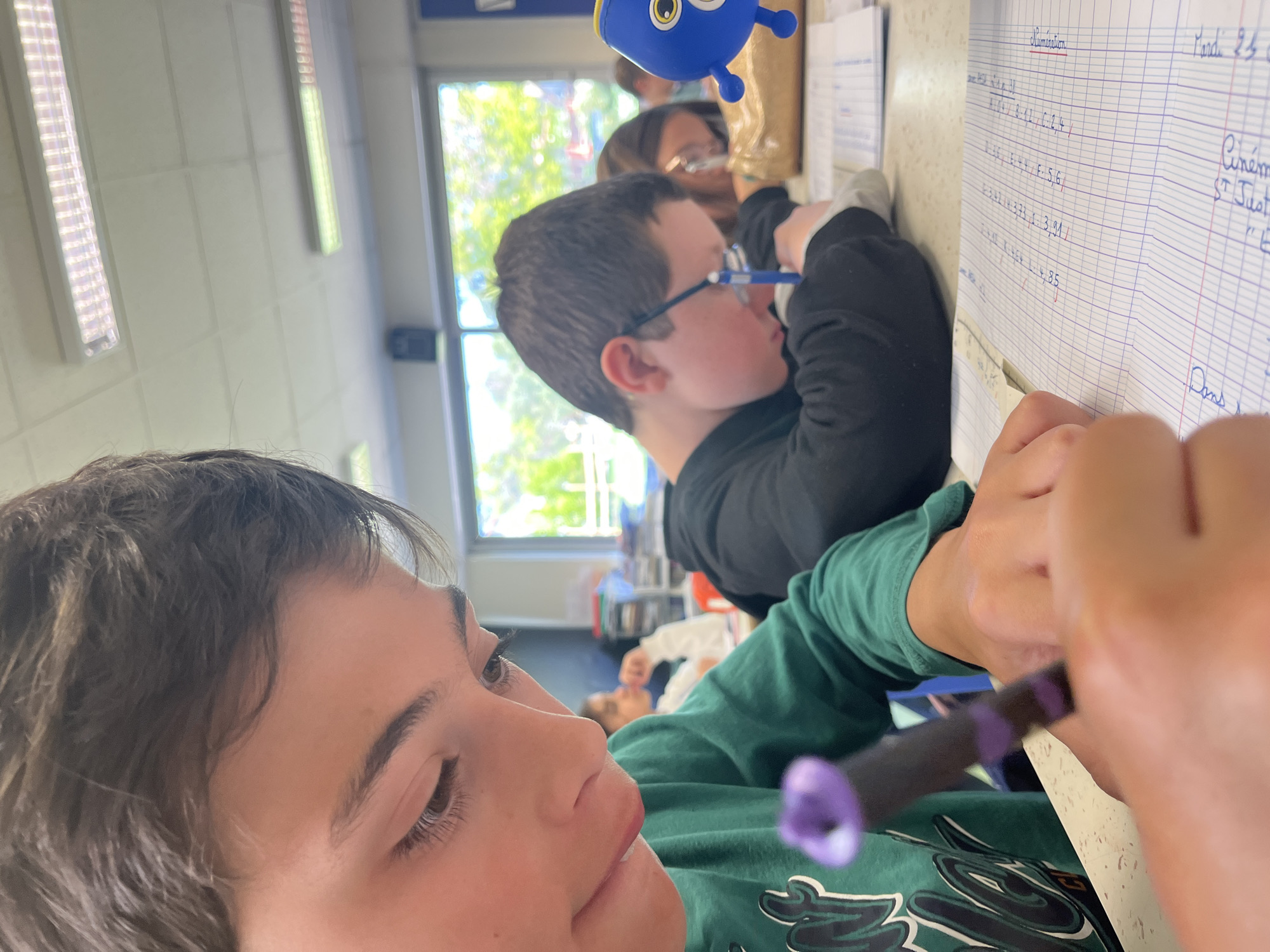 The image, rotated 90 degrees clockwise, captures a classroom or study setting in La Fouillouse, France, during daylight hours.
In the foreground, a young person with dark, wavy hair, wearing a green long-sleeved shirt, is engrossed in writing. They hold a dark pen with a purple tip, poised over a notebook, indicating a moment of concentration on an academic task.
In the midground, another young boy wearing glasses and a black long-sleeved shirt is also focused on his work at a desk. Behind him, a partially visible adult, possibly a teacher, observes the students. A small, blue, cartoonish figure, reminiscent of Dory from "Finding Nemo," is perched near the boy's desk.
The background features a large window showcasing bright natural light and vibrant green foliage outside, affirming it is daytime. Inside, long fluorescent light fixtures illuminate the room. Another individual is partially visible lying on the floor in the deeper background.
The primary activity taking place is classroom learning or homework, with students actively engaged in writing. Visible text on one of the notebooks includes the French heading "Numération" (Numeration) and dates such as "Mardi 23" (Tuesday 23rd). Below this, various mathematical-looking entries are written in blue ink, including sequences like "A: 0,8 / B: 2,1 / C: 2,4 /", "D: 3,6 / E: 4 / F: 5,6", "G: 3,93 / H: 3,91", and "I: 4,59 / J: 4,85". Other partial words like "Cinéma" and "St Just" are also discernible. The scene conveys a focused and productive learning environment.