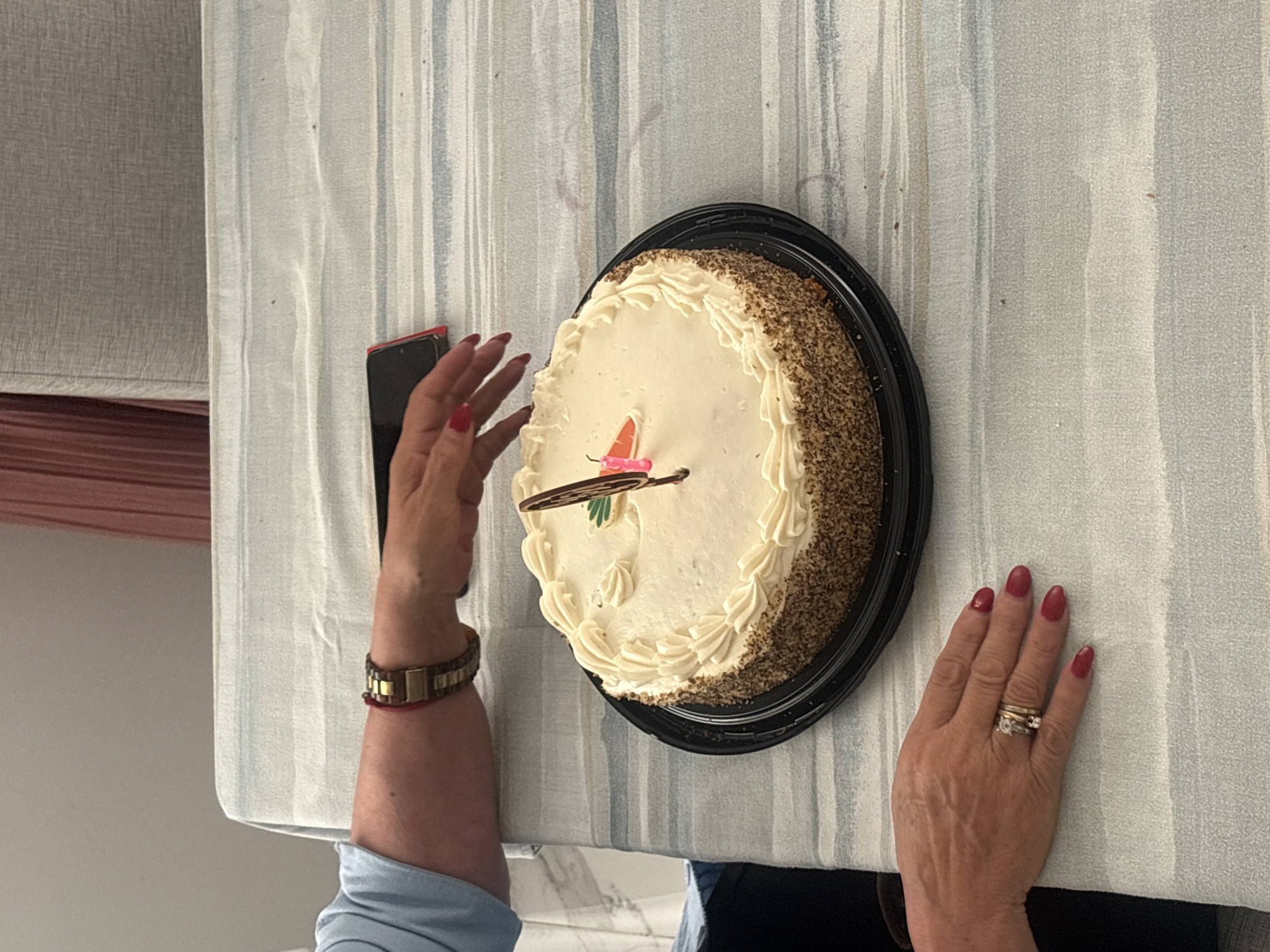 A top-down shot shows a person taking a picture of a frosted carrot cake on a table. The cake is round with white frosting and a decorative carrot on top. The person's left hand, wearing a watch, holds a cell phone, while their right hand, adorned with rings, rests on the table next to the cake. The table is covered with a striped tablecloth. The location is presumably in San Bernardino Tlaxcalancingo, Mexico, though there is no visible evidence of it.