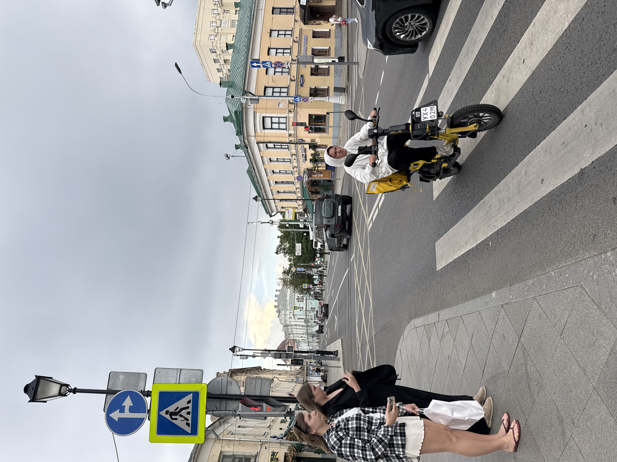 The image depicts a street scene in Moscow, Russia, on a cloudy day. In the foreground, two women stand on a pedestrian sidewalk, one in a black blazer and the other in a plaid shirt and white skirt. A person on a yellow electric scooter, wearing a white hoodie and black pants, is crossing a pedestrian crosswalk. The scooter has a yellow delivery bag attached to the back and a license plate that reads "YX4 02M". In the background, cars are visible on the street, and various buildings line the street, including a yellow building with green trim. Traffic signs and streetlights are also present.