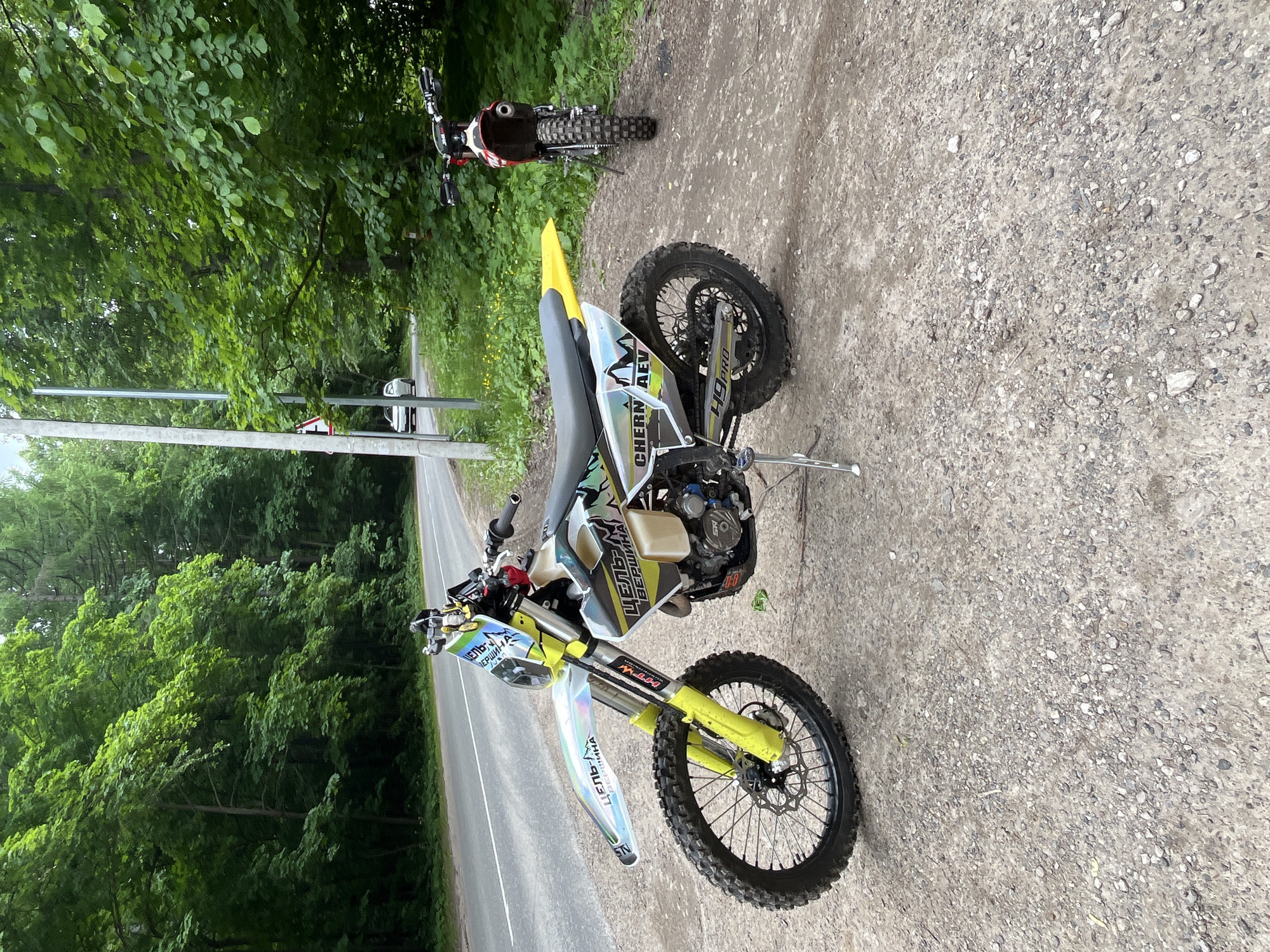 The image depicts two off-road motorcycles parked on a gravel and dirt shoulder beside an asphalt road in a forested area, identified as Ореховка, Russia. In the foreground, positioned to the left, is a white, yellow, and black enduro motorcycle with knobby tires, resting on a kickstand. Its graphics include "ЦЕЛЬ-ВЕРШИНА" (Target-Summit/Peak) on the front fender and side panel, and "CHERN AEV" and "H9 PRO" on the rear sections. Its front forks and rear fender are yellow. Further back and to the right, a second, red and white enduro motorcycle with knobby tires is parked, facing away from the camera.
The asphalt road runs from left to right, bordered by a solid white line, and curves gently to the left in the distance. A tall, slender pole, possibly a light pole, stands near the road's edge, alongside a red and white triangular warning sign. Dense green foliage and tall trees line both sides of the road, forming a thick forest canopy. In the background, on the road, a white car is partially visible, moving away from the viewer. The ground on the shoulder is a mix of small gravel, dirt, and sparse green vegetation with a few yellow wildflowers. The sky is overcast. No people are present in the scene.
