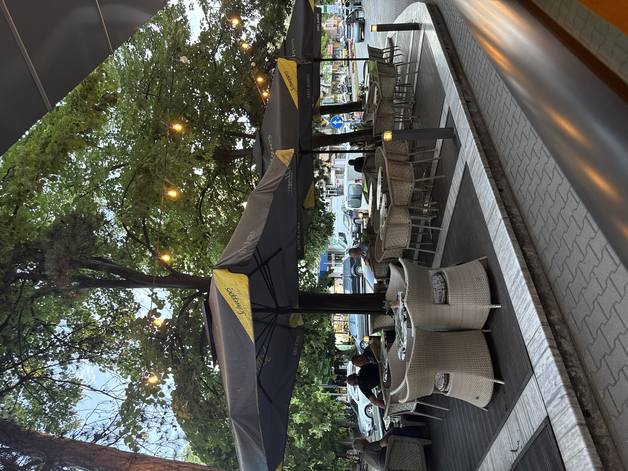 The image depicts an outdoor seating area of a cafe in Stara Zagora, Bulgaria. The cafe has several tables and chairs, arranged under large, black umbrellas with yellow Schweppes branding. People are seated at some of the tables, engaged in conversation or observed by others. The seating area is located on a paved surface next to a sidewalk, and the area is surrounded by trees with string lights. In the background, there is a street with cars and other buildings, suggesting the cafe is located in a commercial area. There is a road sign indicating direction and pedestrian crossing.