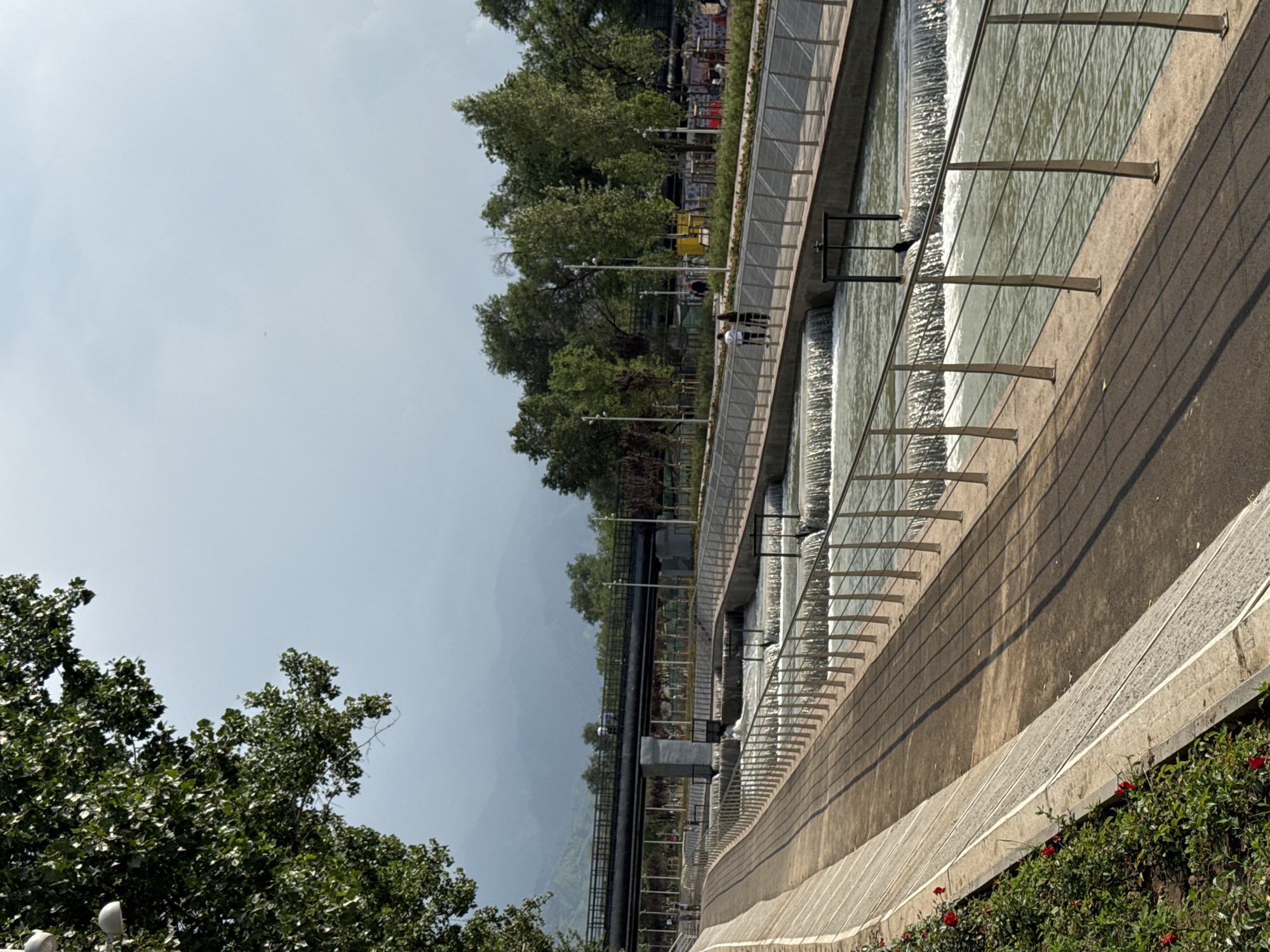 The image depicts a scenic view of the Big Almaty Canal in Almaty, Kazakhstan, on a cloudy day. The canal features a series of cascading waterfalls and is lined with concrete embankments and metal railings. A paved walkway runs along the left side of the canal, bordered by lush green bushes with red flowers. On the right side of the canal, another walkway is visible, with several people strolling along it. In the background, a bridge spans across the canal, with trees and distant mountains under a hazy sky. The scene conveys a sense of urban recreation and natural beauty.